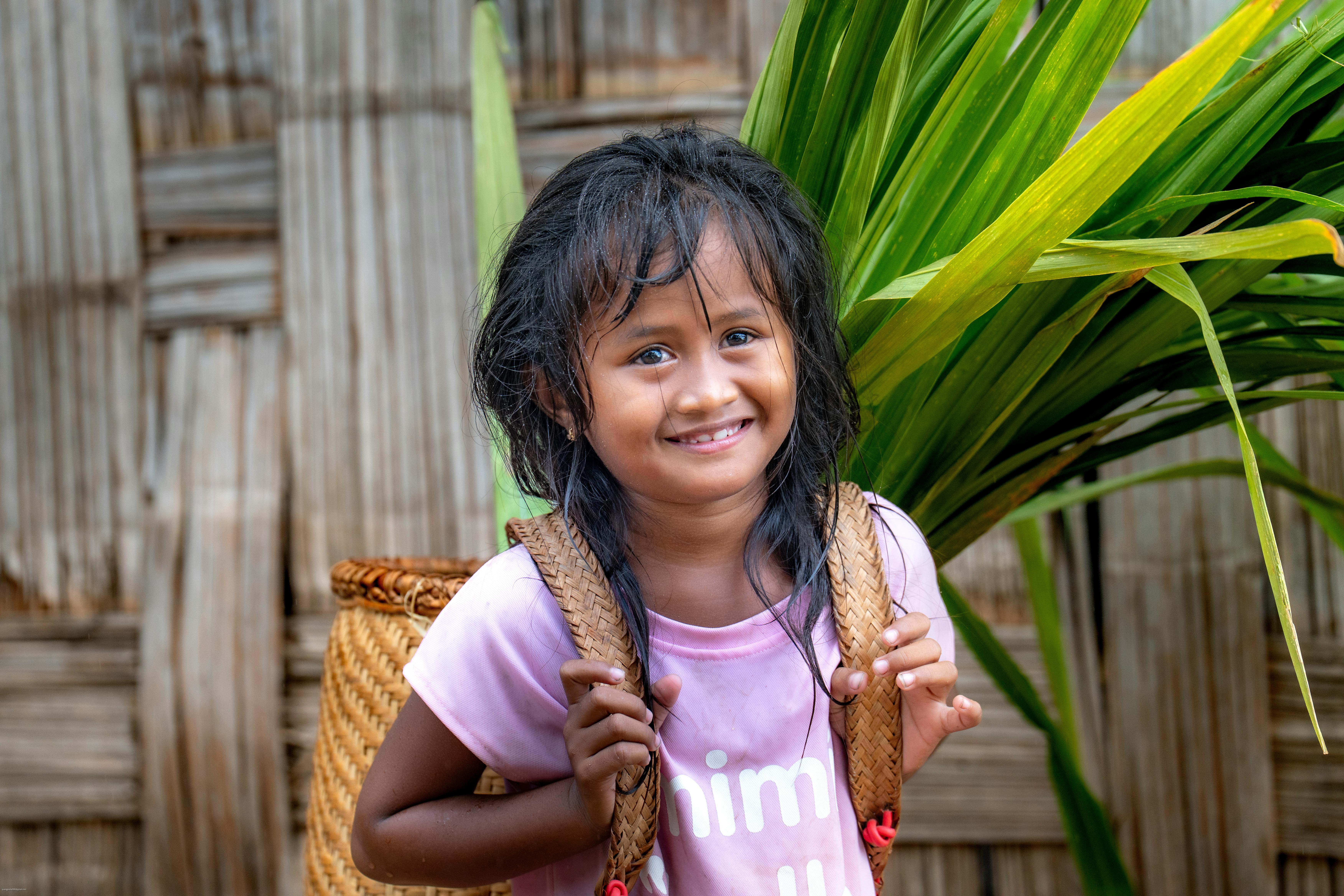 A joyful child with long dark hair smiling in front of tropical leaves, carrying a backpack.