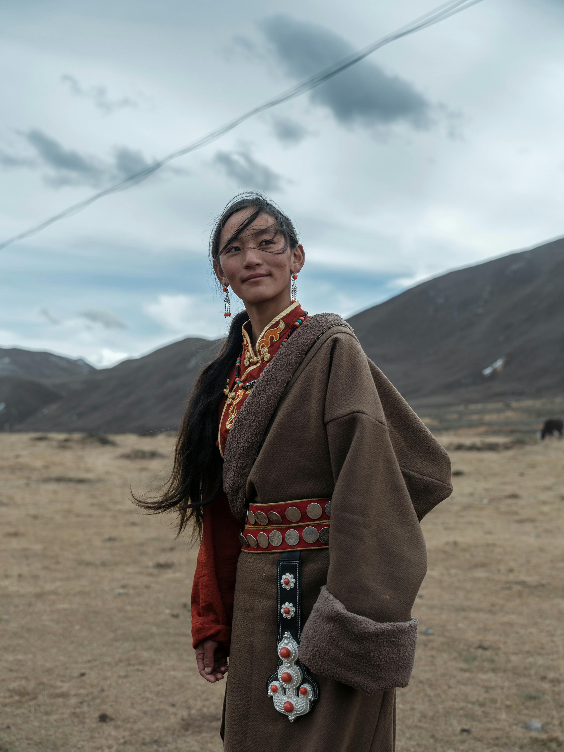 A young woman stands in a Tibetan landscape wearing traditional attire with decorative details.