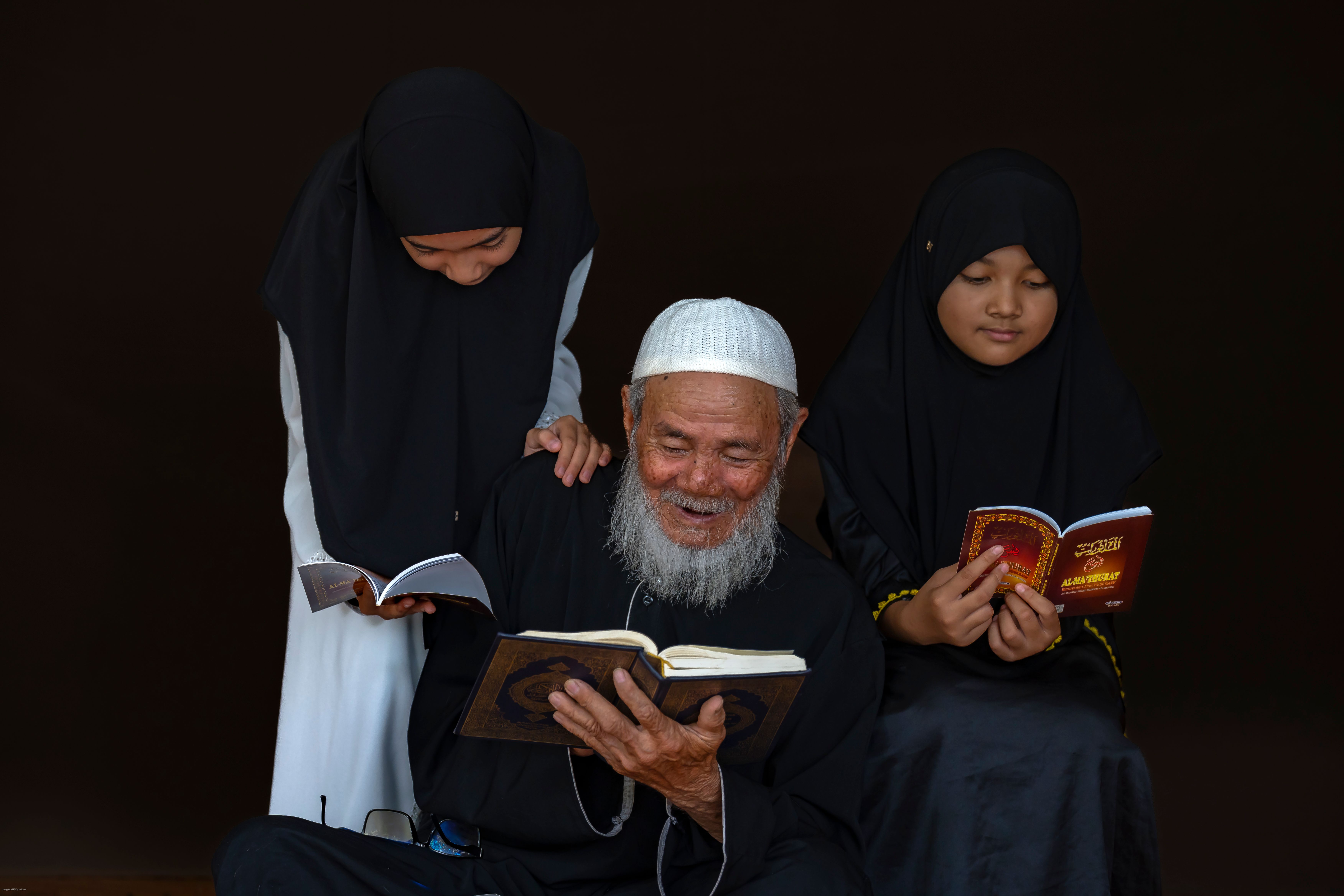 A senior man and two girls in traditional clothing engage in reading together indoors.