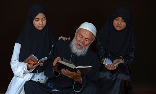 Free An elderly man reading a book with two young girls in traditional attire, fostering education and tradition. Stock Photo