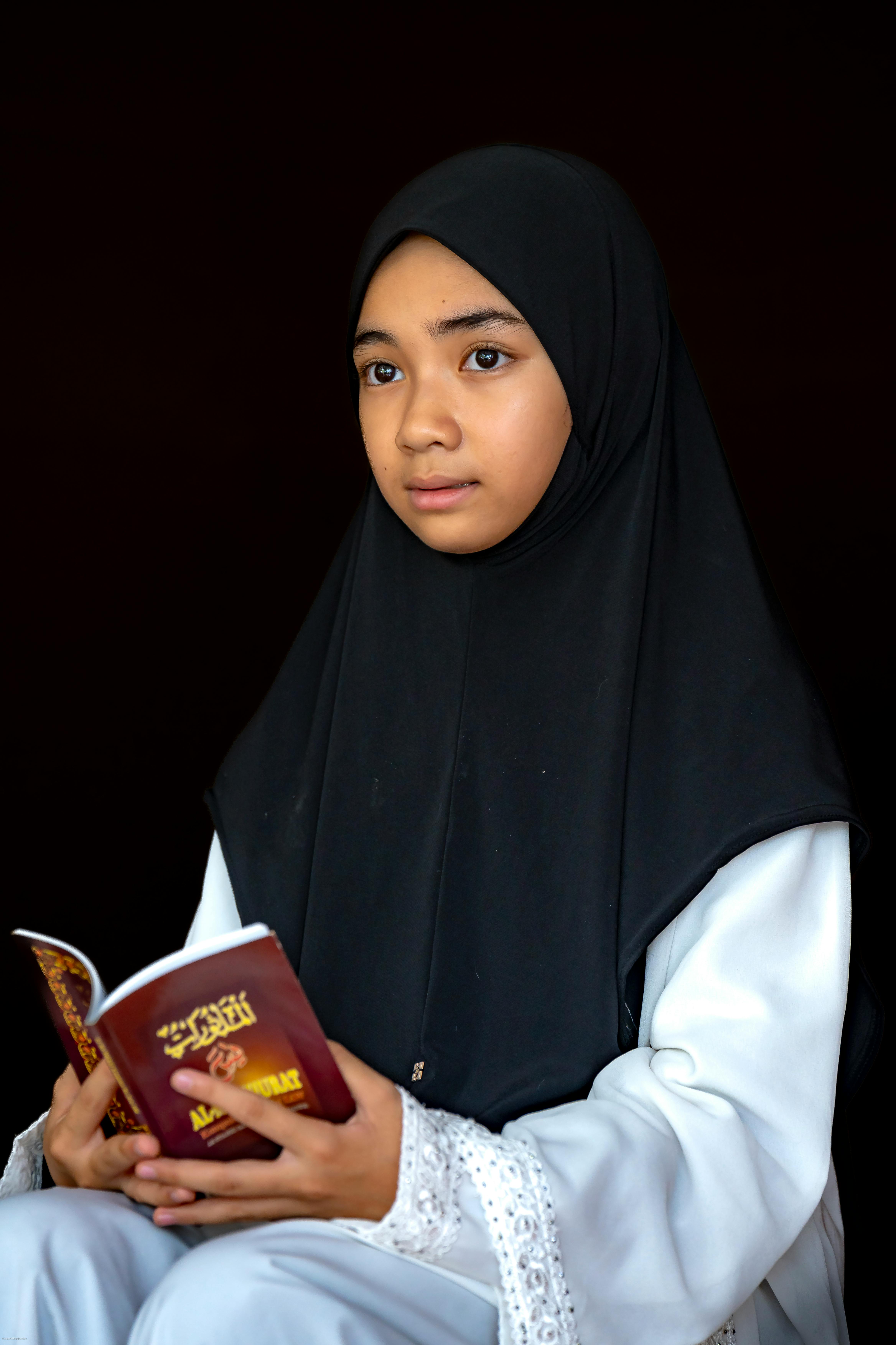 A young girl in a hijab attentively reading a book on a dark background, depicting cultural and religious themes.