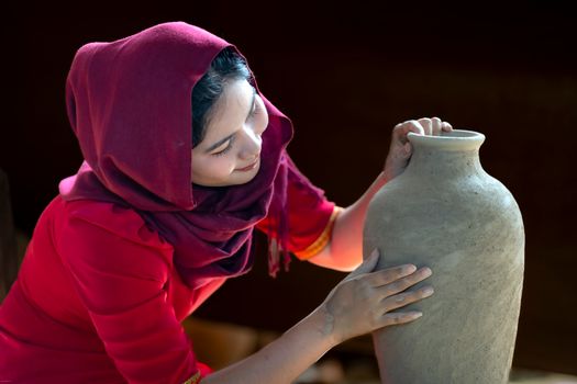 A young woman in a red headscarf attentively sculpts a large clay vase indoors.