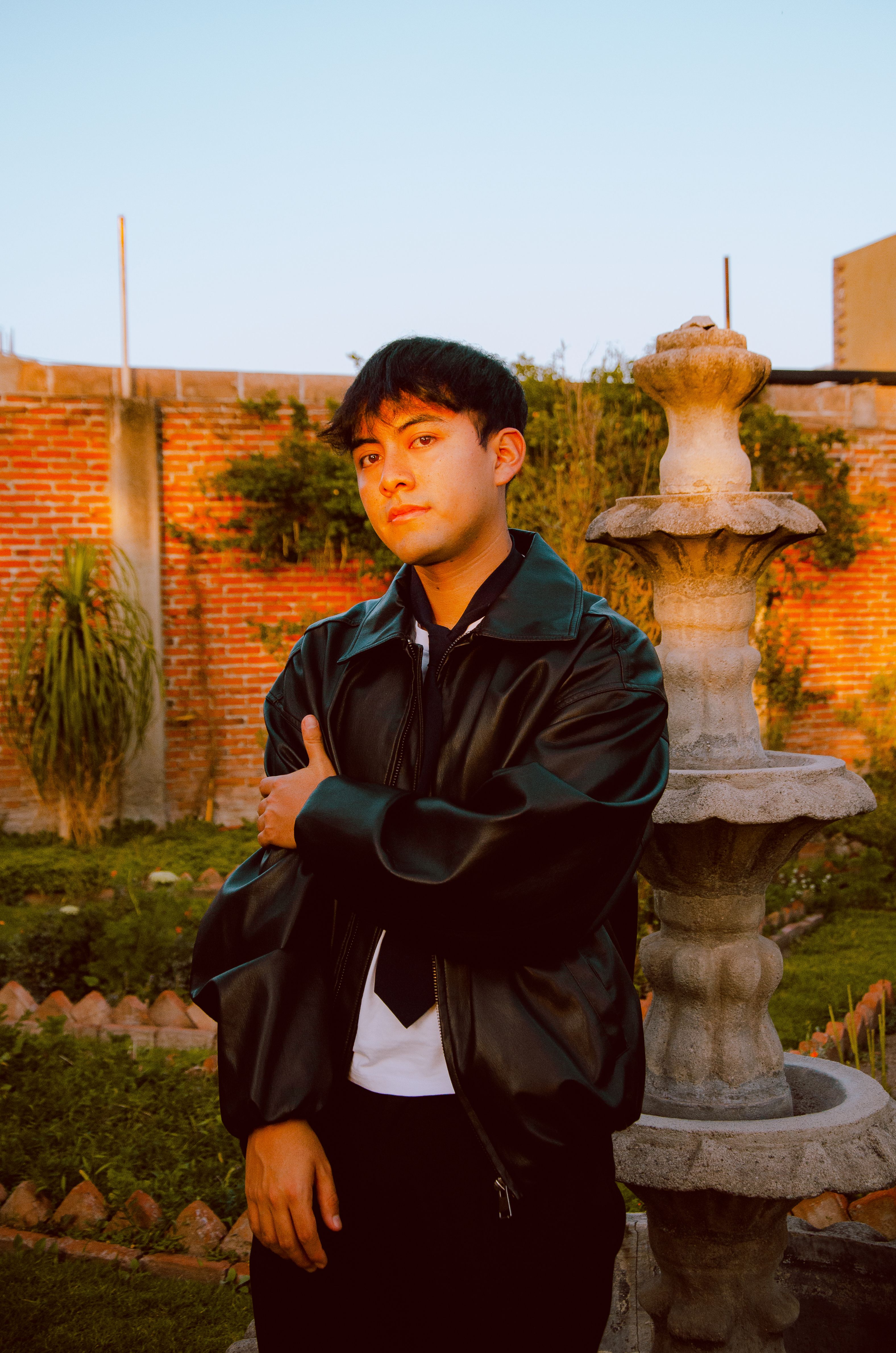 An adult male in a leather jacket poses by a stone fountain in a backyard garden with warm sunlight.