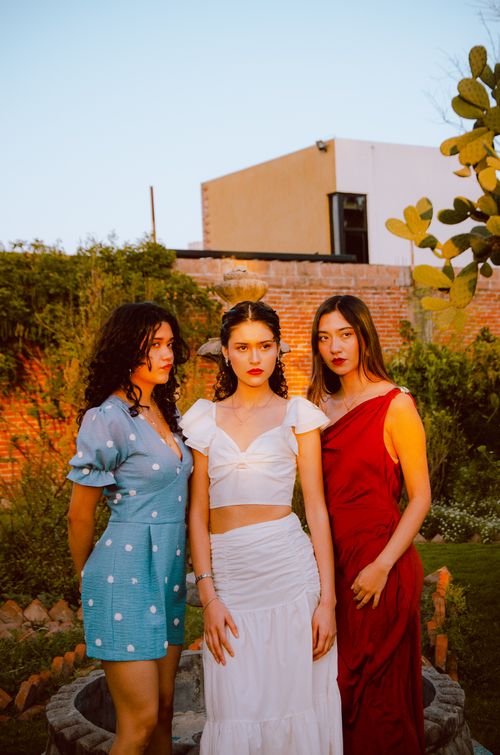 Free Three women pose stylishly in a sunlit garden setting, showcasing vibrant fashion attire. Stock Photo