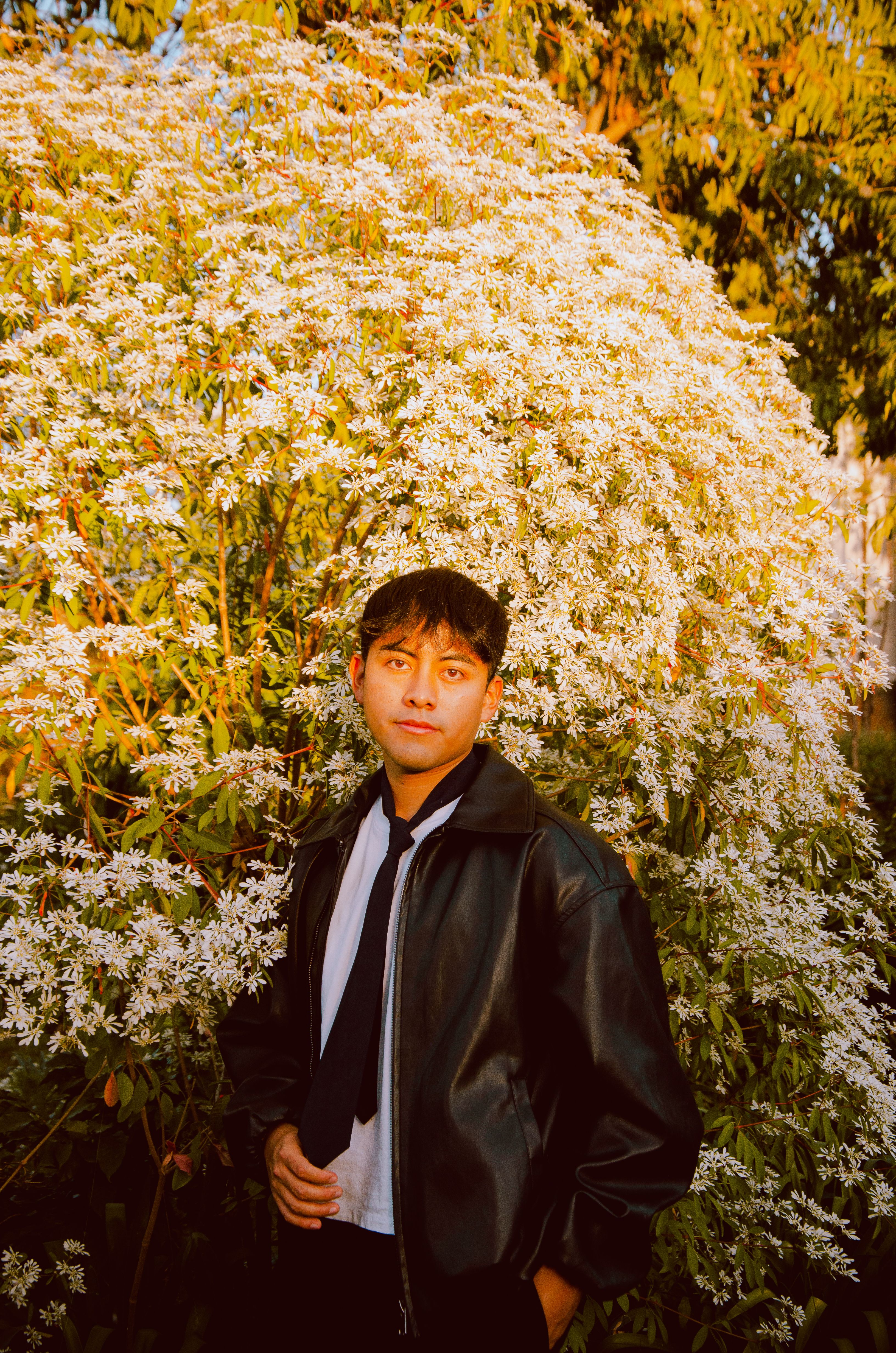 A young man poses in front of a flowering bush, exuding confidence and style.