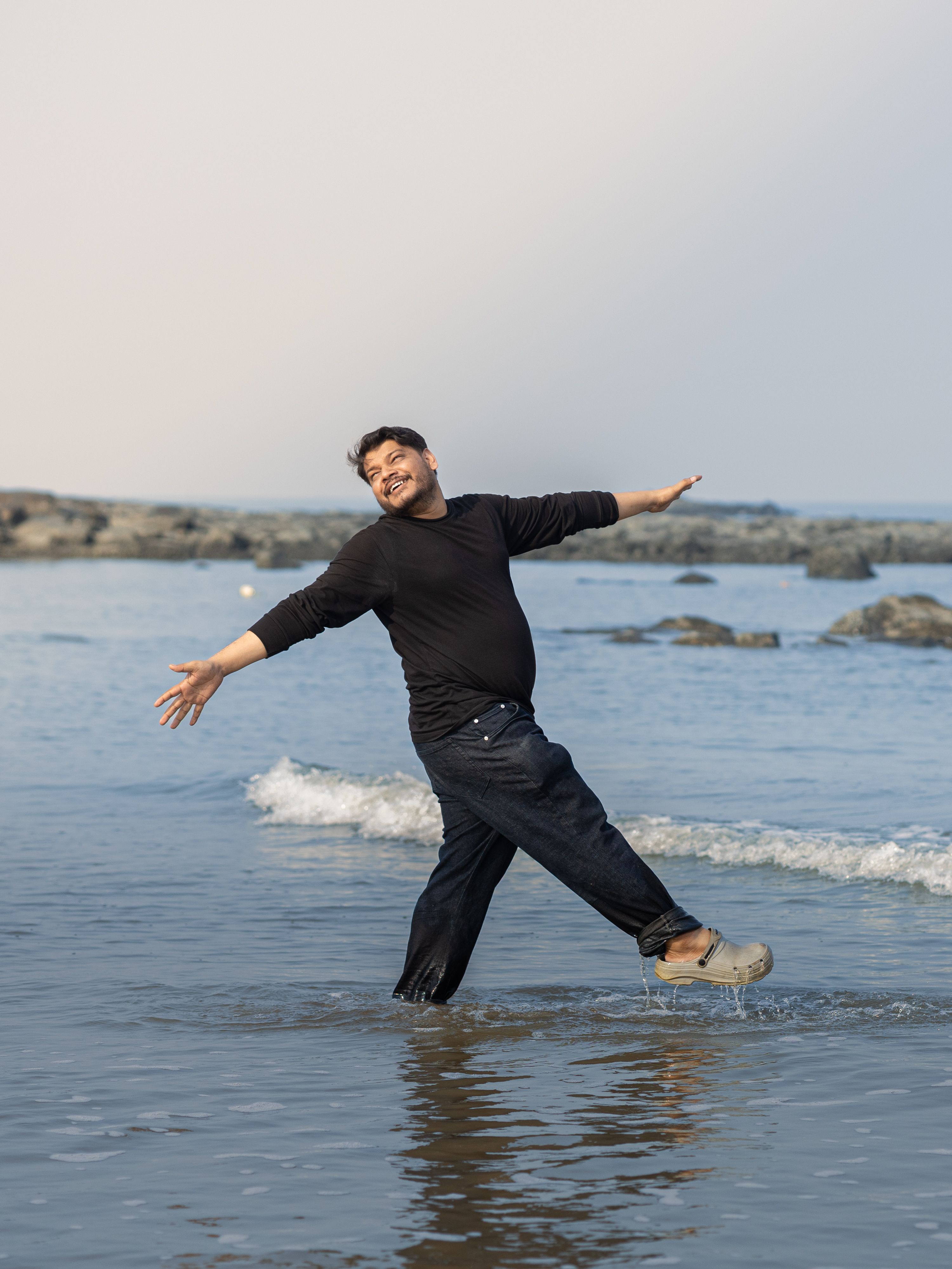 A smiling man in casual attire joyfully dances in shallow ocean water on a sunny day.