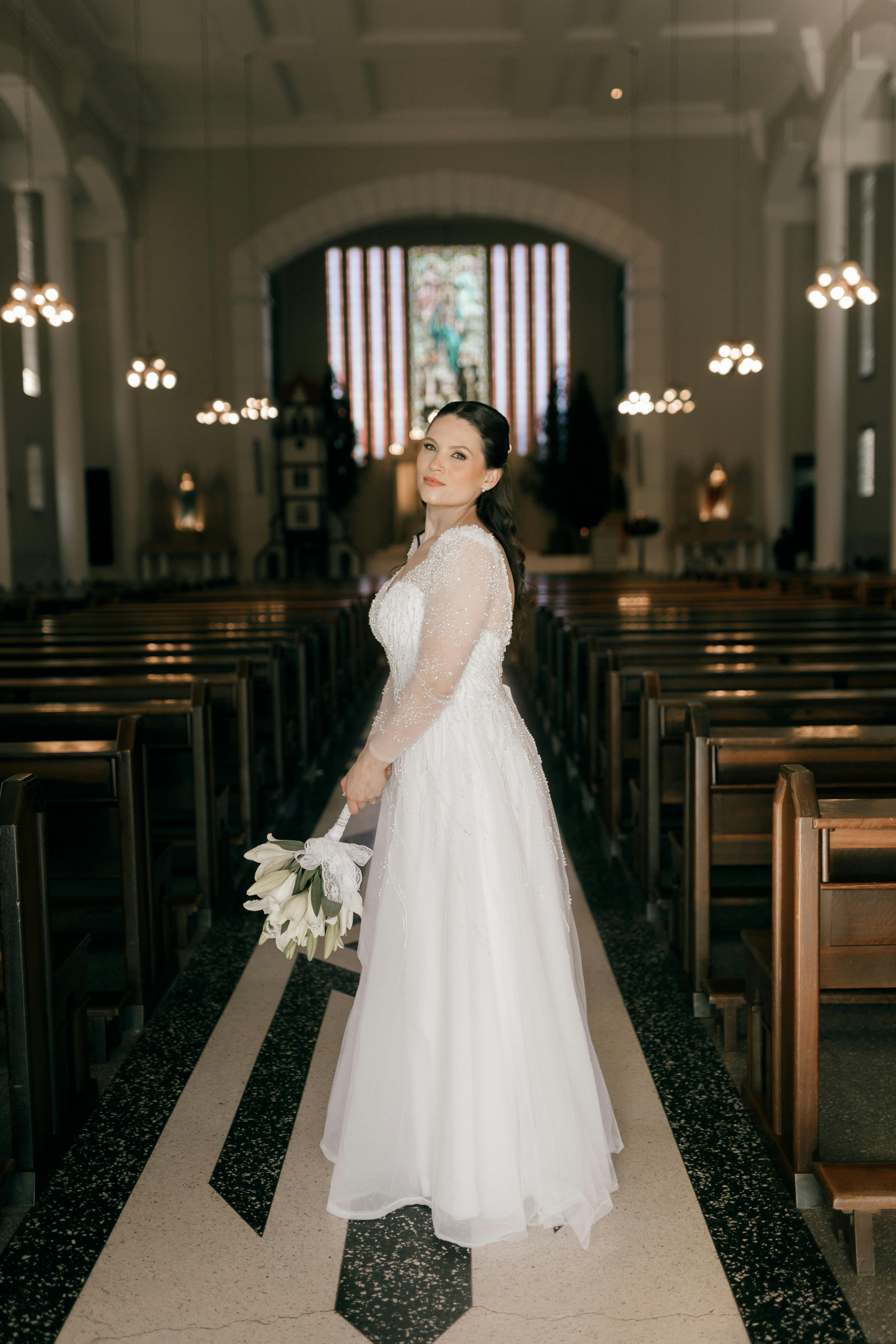 Bride in a white gown holding flowers inside a beautifully lit church.