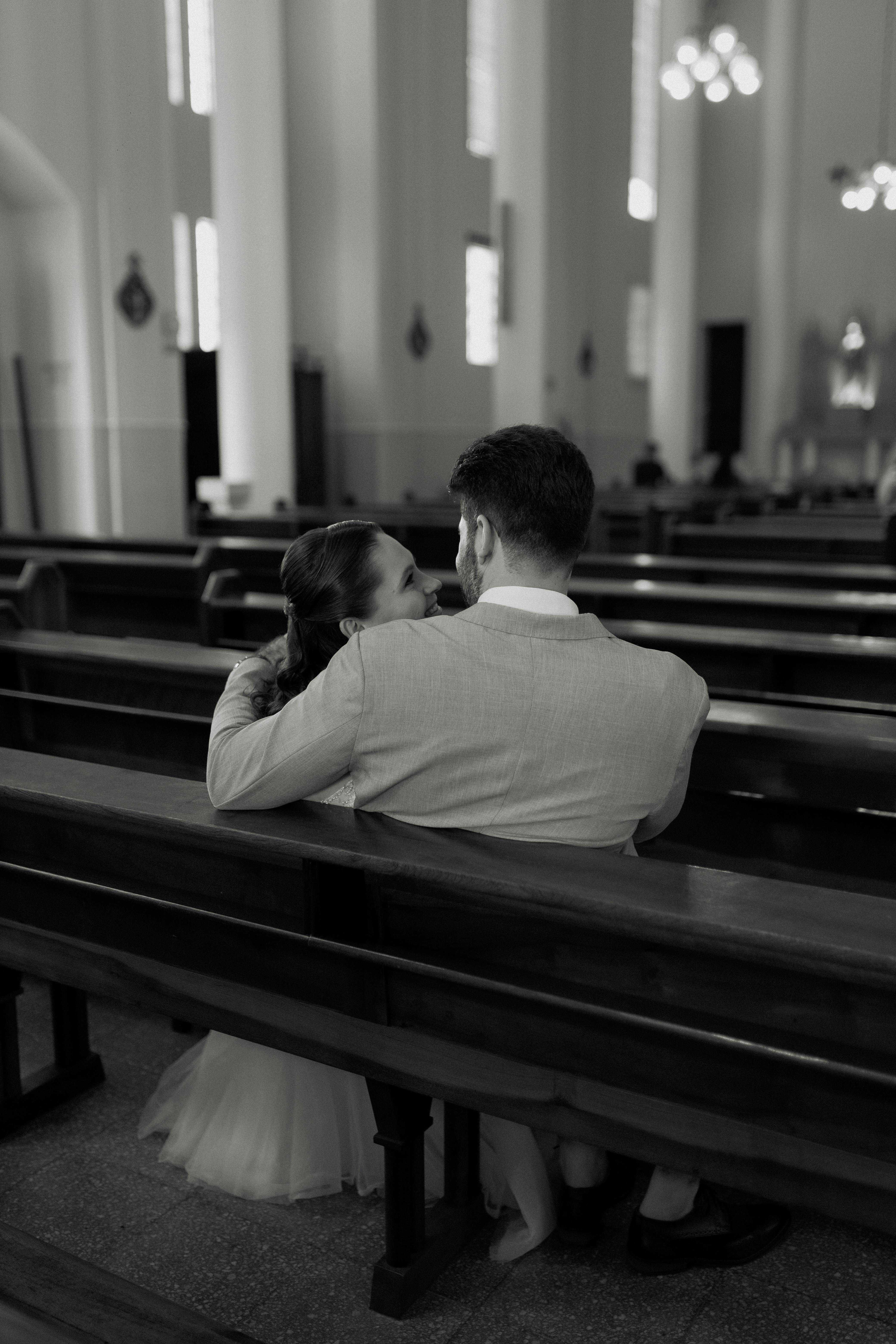 A couple in a wedding dress and suit sit closely on a church pew, sharing a tender moment.