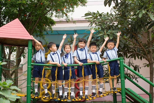 Group of smiling schoolchildren in uniforms raising hands on a playground bridge outdoors.