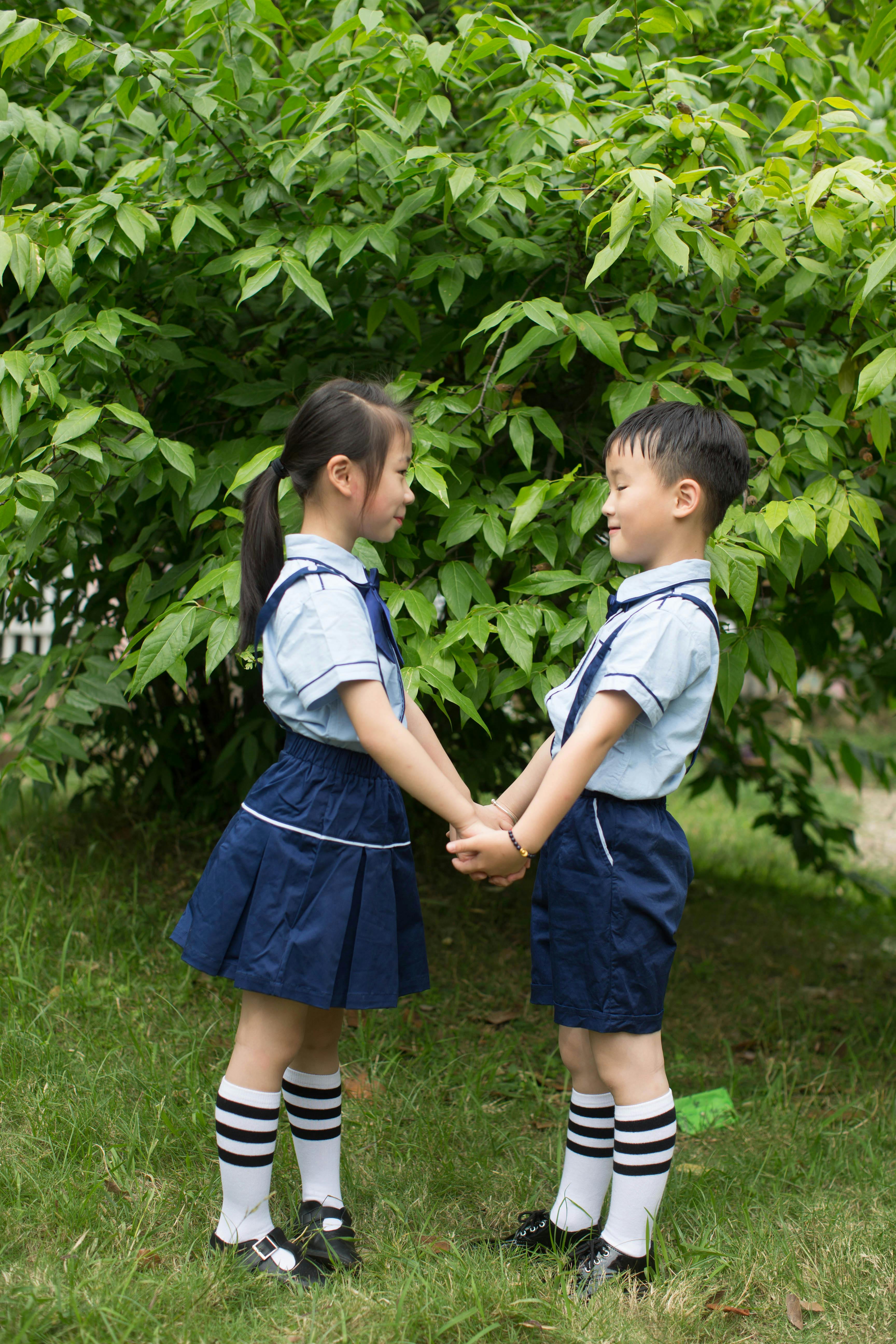 Two young children in school uniforms holding hands against a leafy green background.
