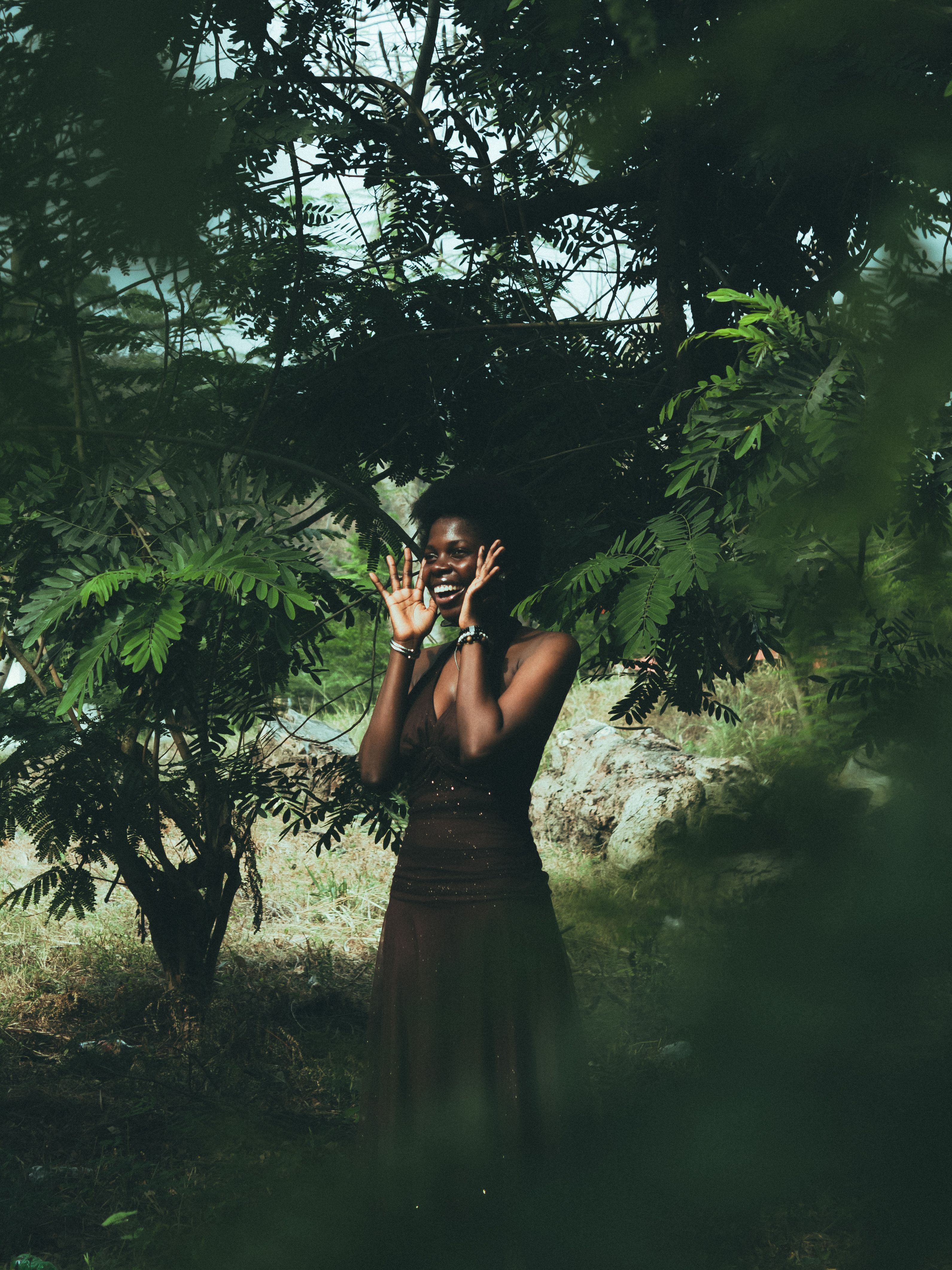 A woman joyfully poses amidst lush green trees, expressing happiness.
