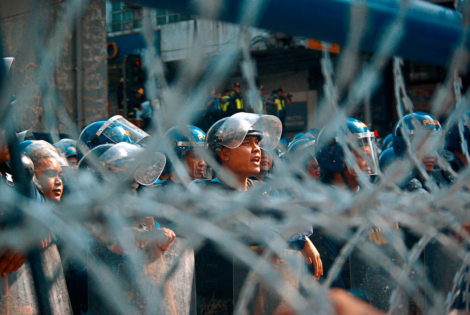 Riot police lined up behind barbed wire during a protest, showing tension and security forces.