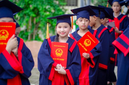 Group of children in graduation gowns holding diplomas outdoors in Hà Nội, Việt Nam.