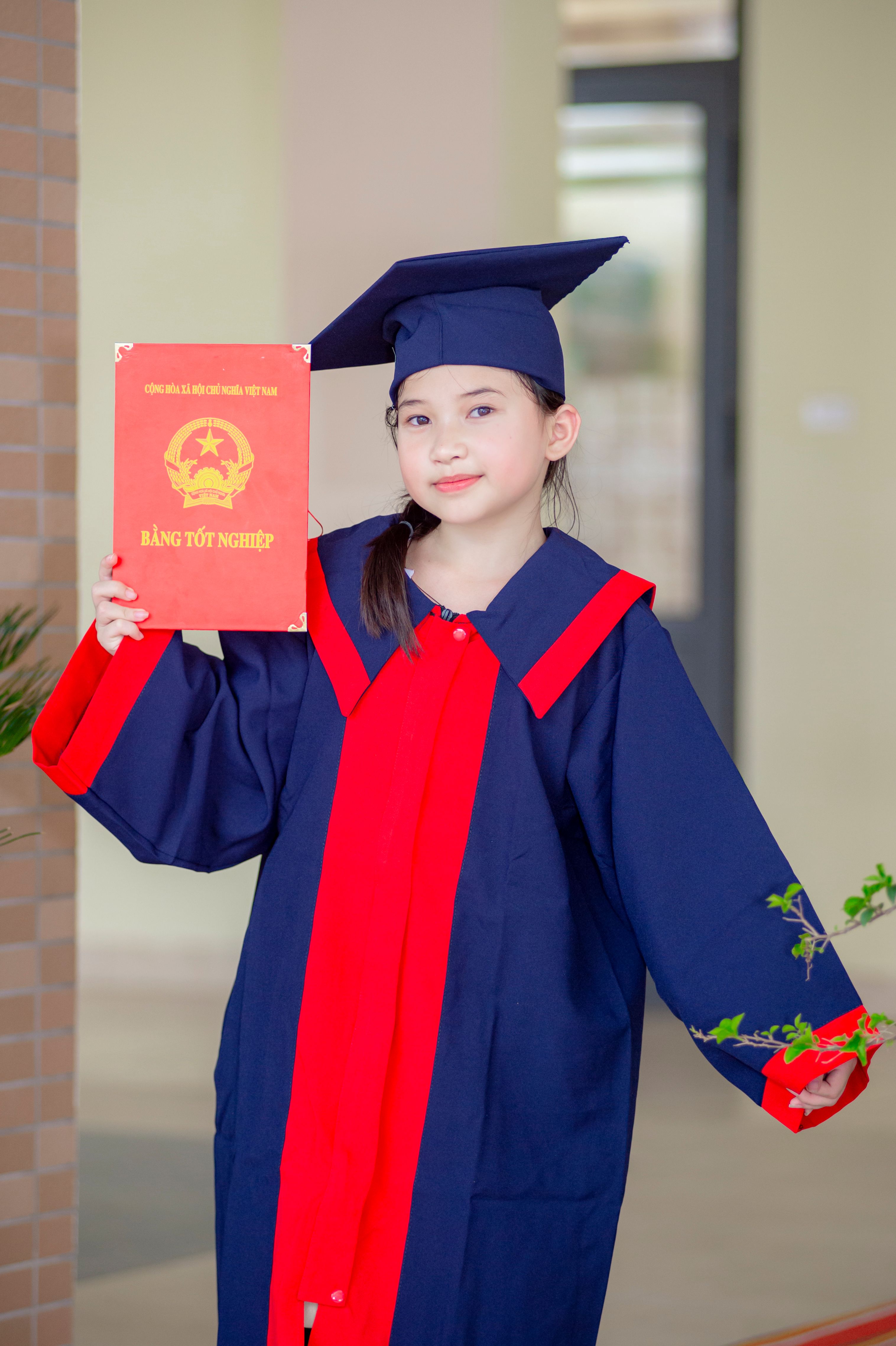 A young girl in graduation outfit holding a diploma, celebrating commencement in Hanoi, Vietnam.