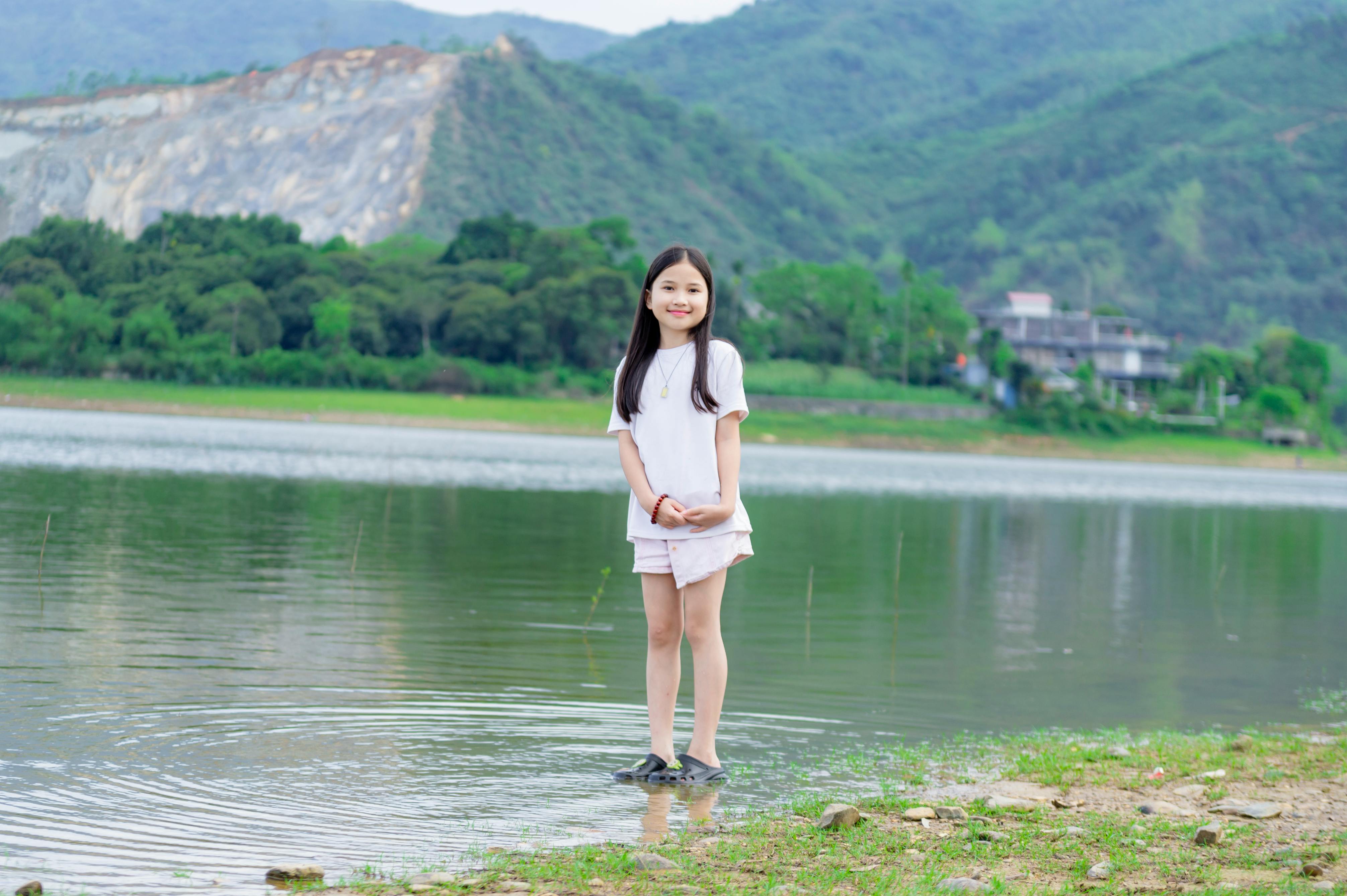 Free A young girl stands by a tranquil lake with scenic mountains in Hanoi, Vietnam. Stock Photo
