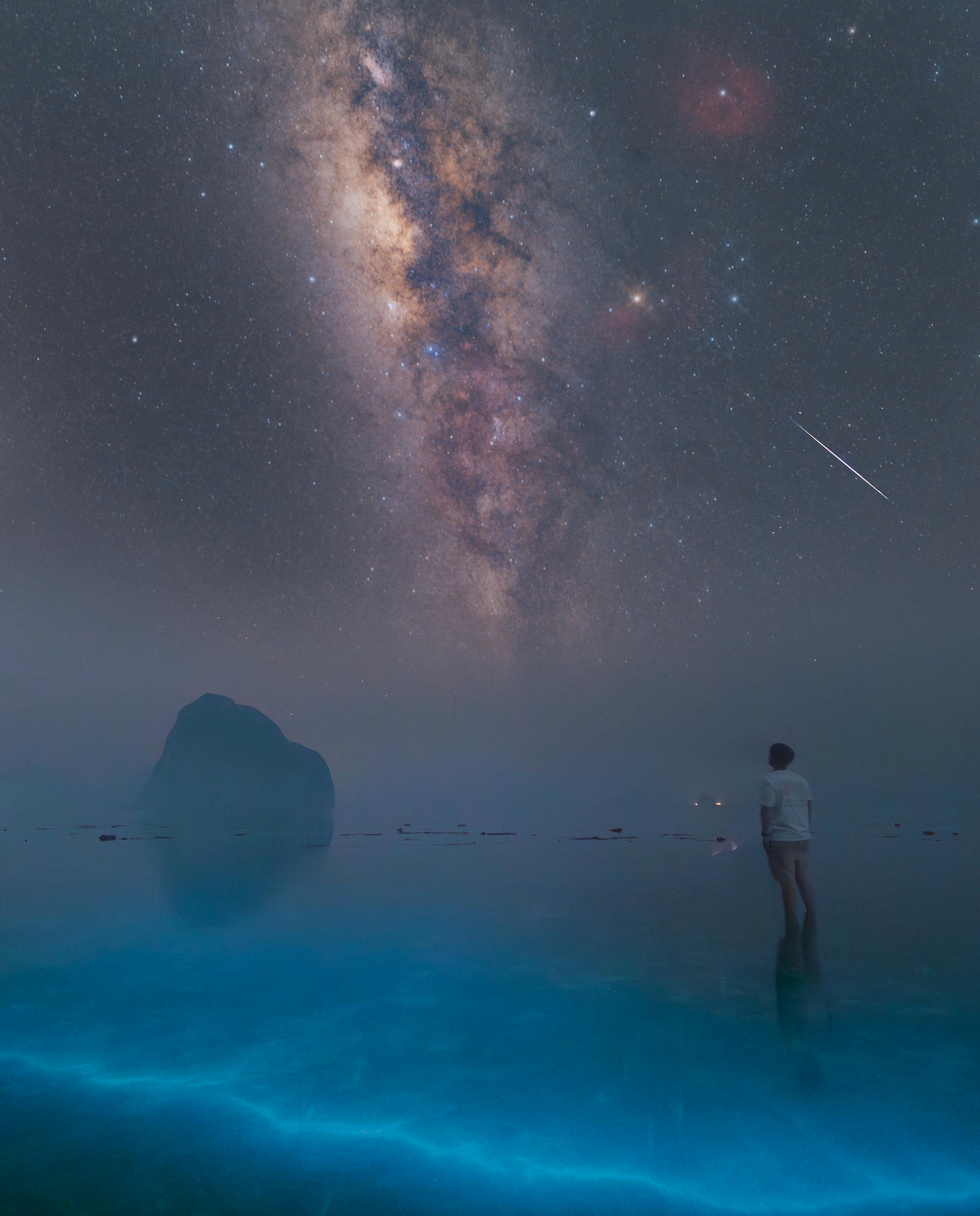 A person gazes at the Milky Way under a starry sky by the illuminated ocean.