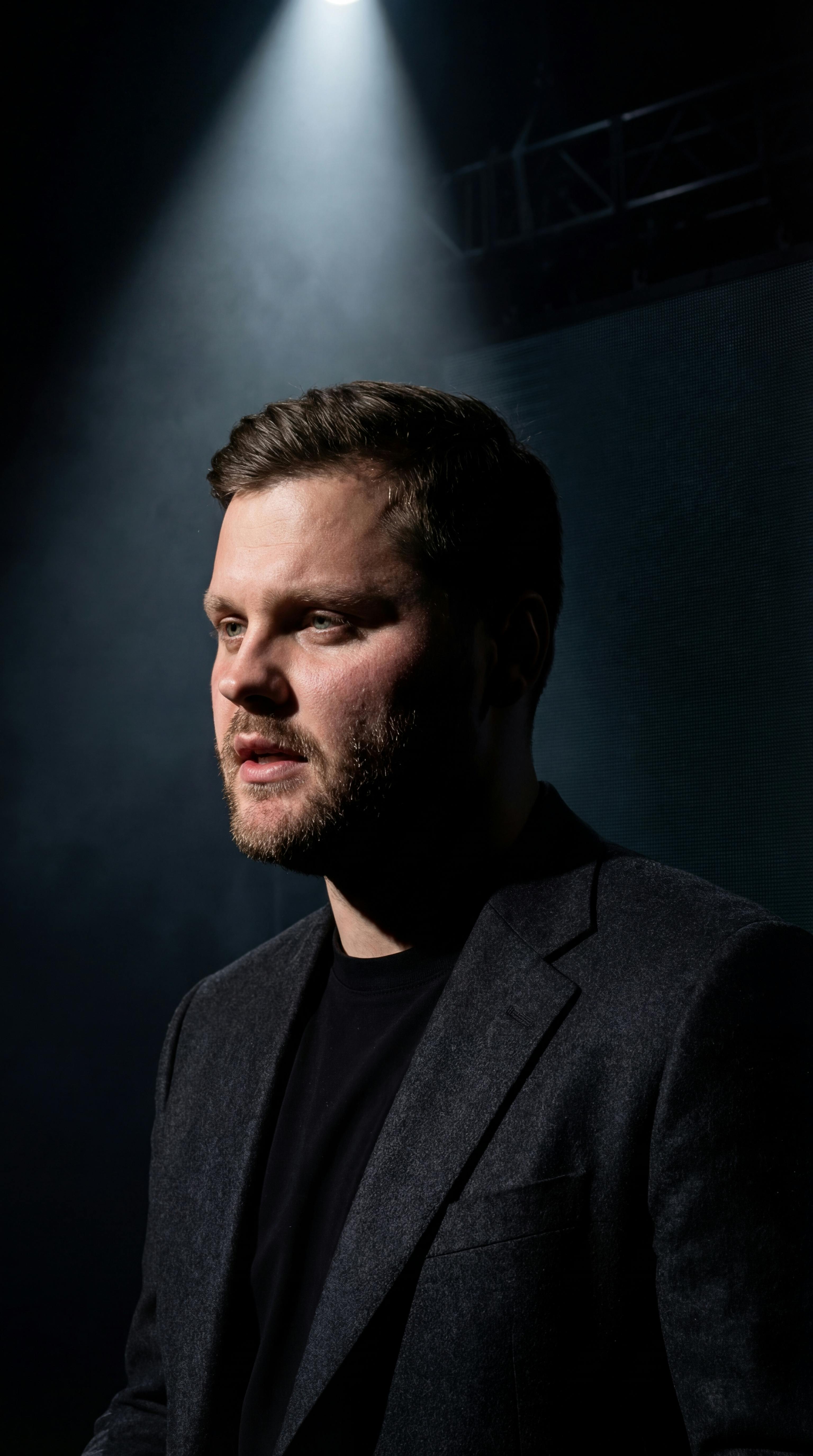 免費 一束聚光燈照亮了一位身穿深色西裝的成年男子，畫面極具戲劇性。 圖庫相片