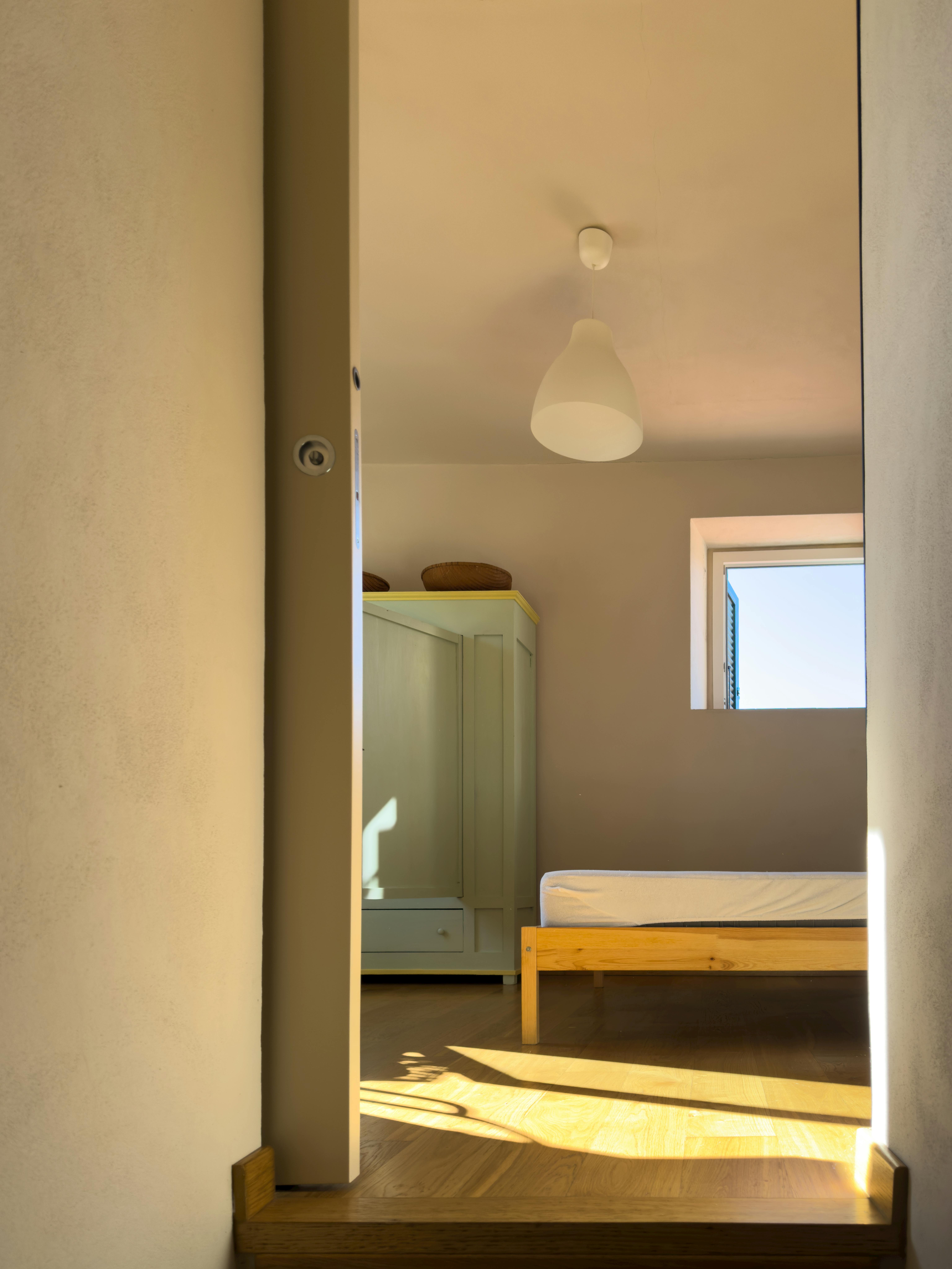 Simple minimalist bedroom with wooden bed and wardrobe, bathed in natural daylight.