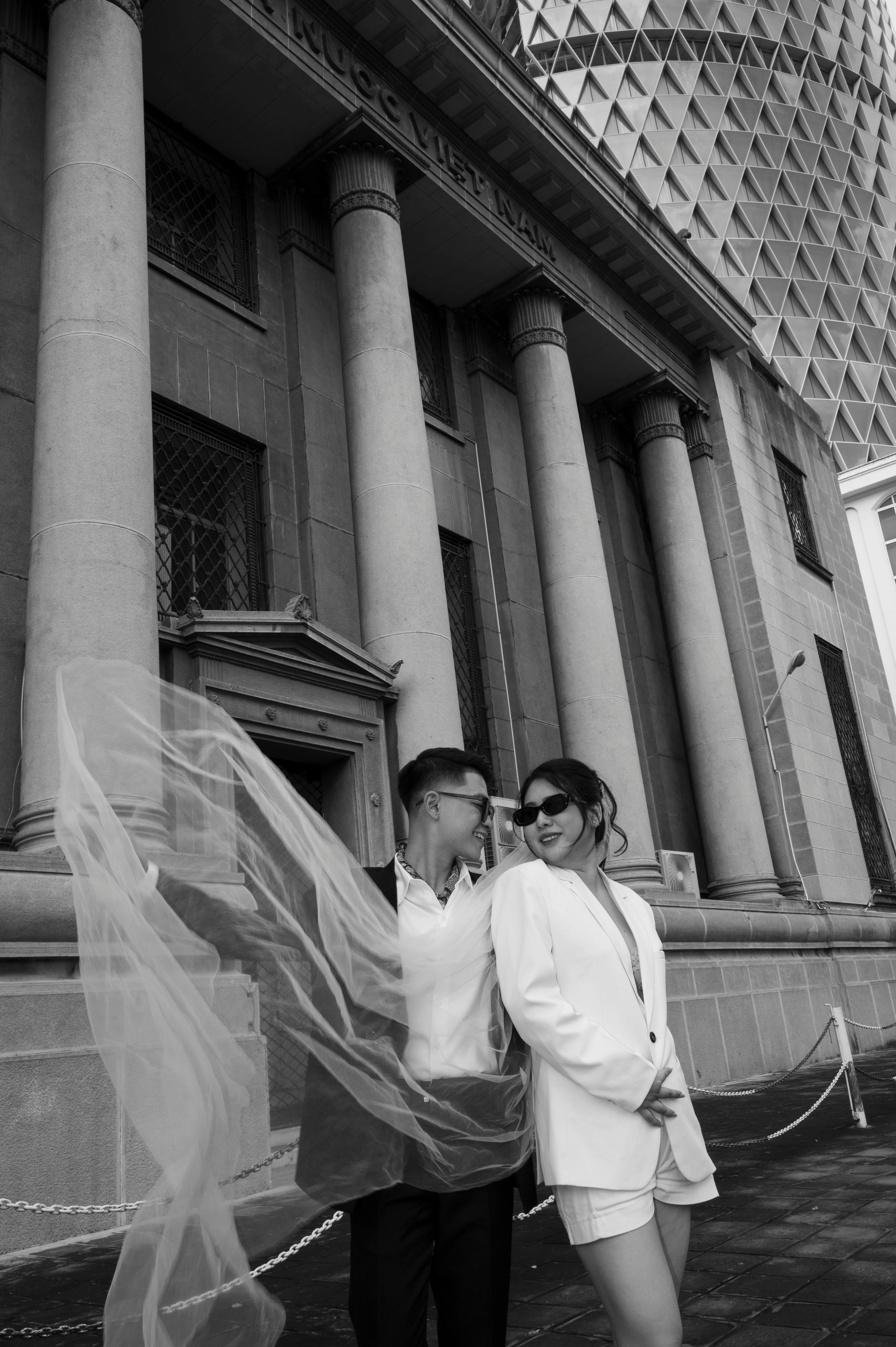 Elegant couple posing with flowing veil in front of architectural columns, urban style.