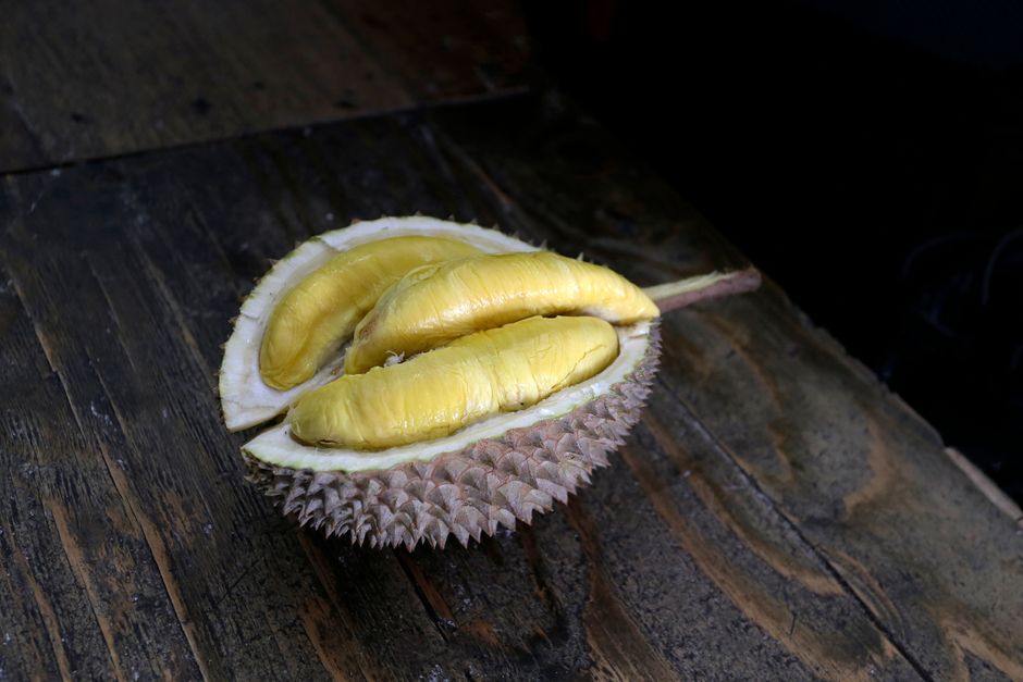 A halved durian showing pale yellow custard-like flesh segments nestled inside the spiky green husk, with whole spiky durians in the background.