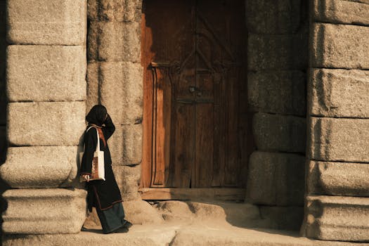 Woman in traditional attire stands beside a historic stone doorway under warm daylight.