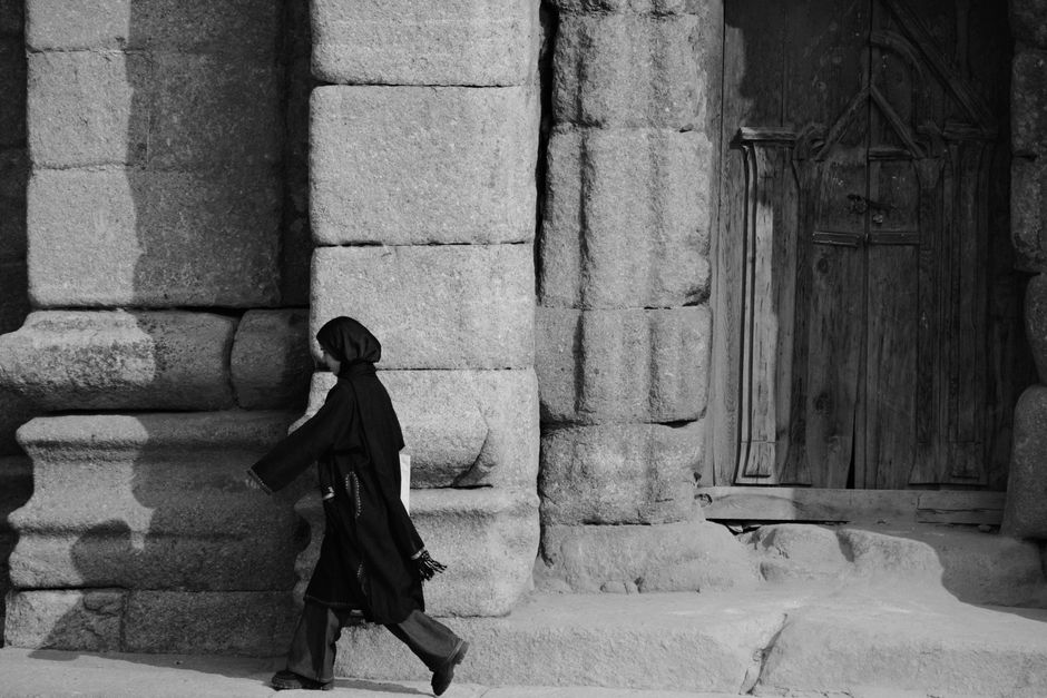 A person in traditional attire walks past an ancient stone wall and wooden door in black and white.