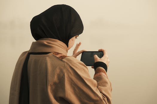 A woman wearing a hijab captures a photo with her smartphone, showcasing modern lifestyle.