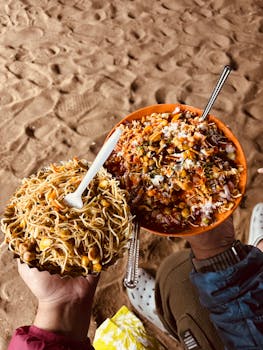 Vibrant street food bowls of chaat and noodles held over sandy beach in Puri, India.