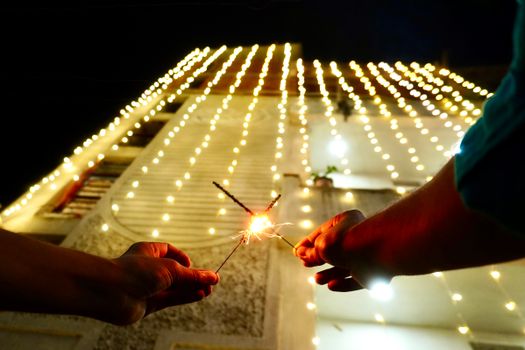 Hands holding sparklers in front of a brightly lit building during Diwali festivities at night.