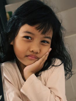 A young girl indoors with a thoughtful expression resting her face on her hand.