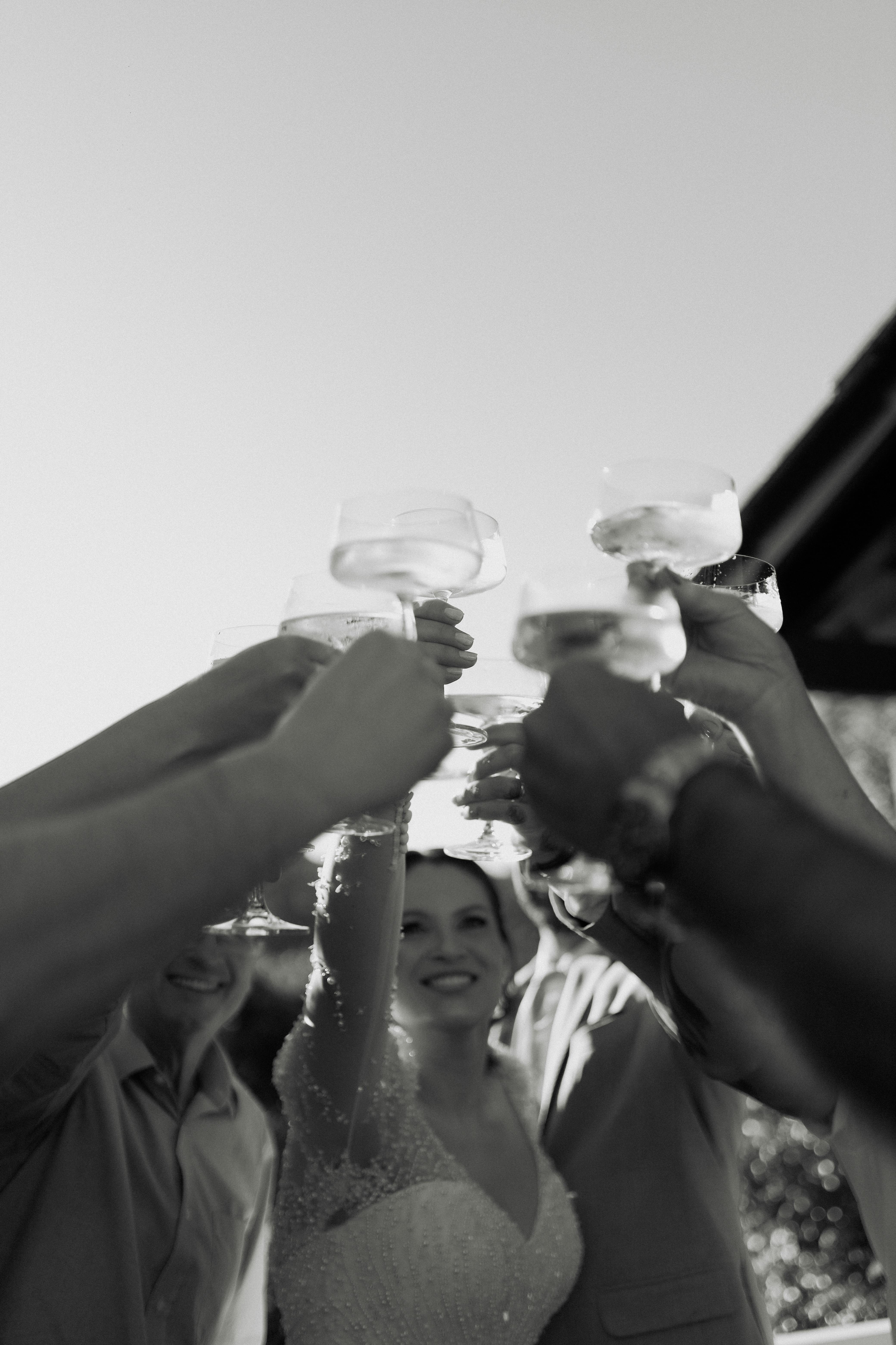 Black and white photo of a joyful wedding toast with friends raising glasses outdoors.