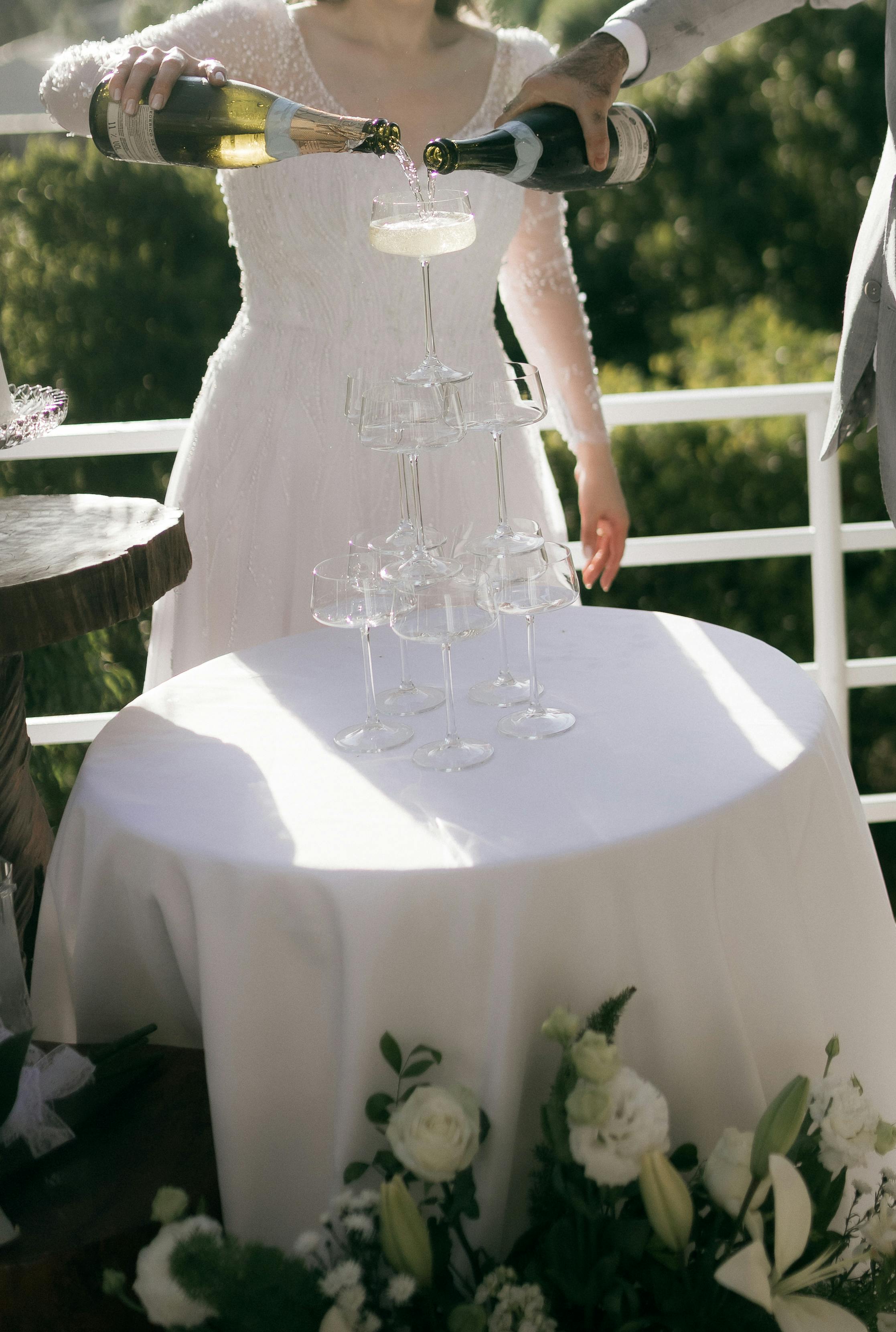 Bride and groom pouring champagne into stacked glasses outdoors at a wedding.