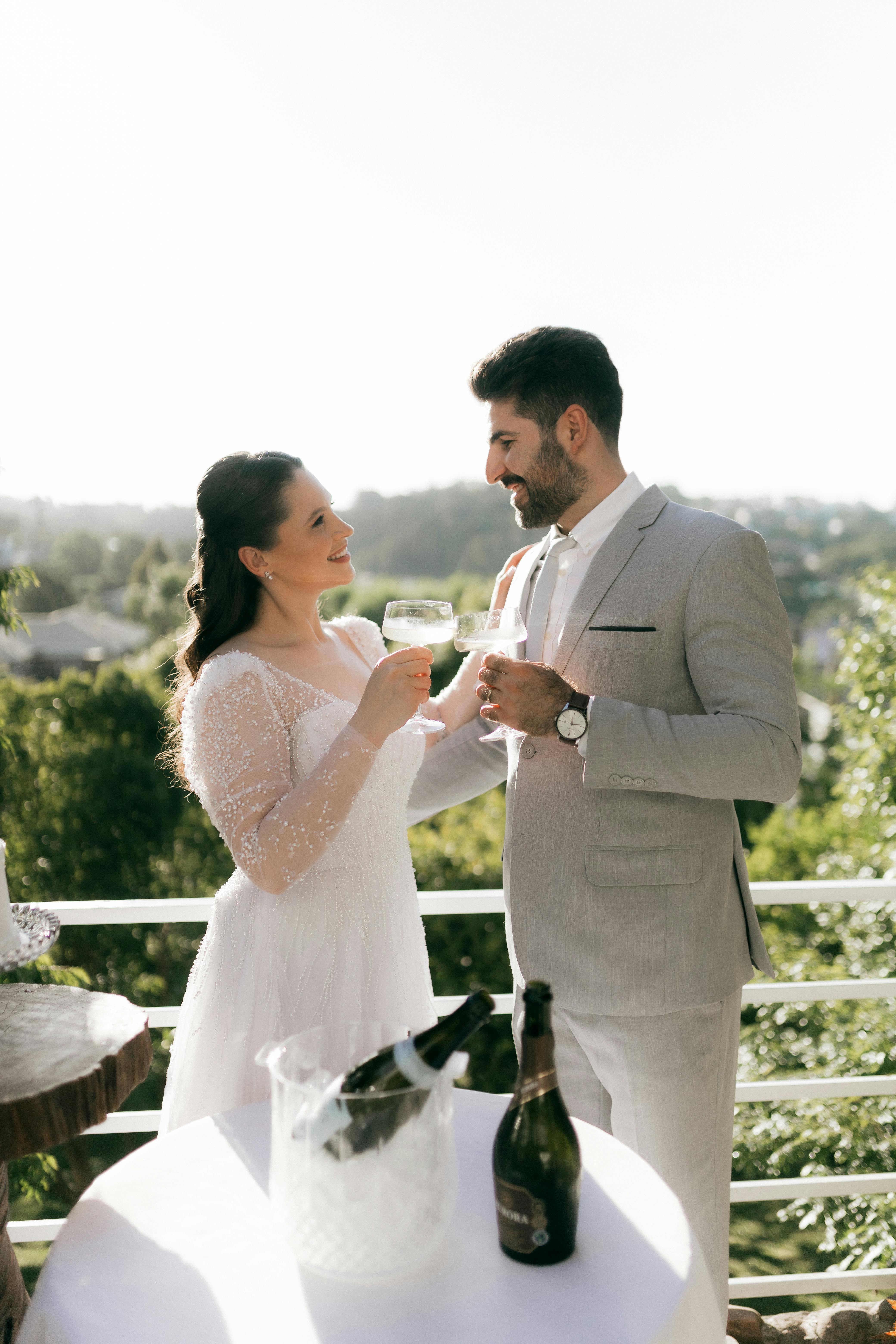 A couple in wedding attire toasting with champagne on a sunny day outdoors.