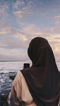 A woman in a hijab stands by the ocean, taking a photo with her mobile phone during sunset.