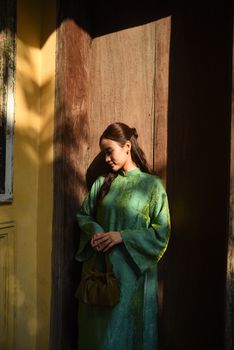 A woman stands in sunlight wearing a traditional green dress, holding a purse.