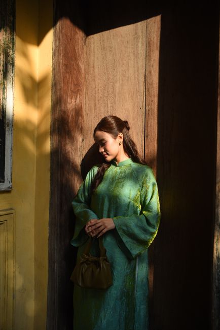 A woman stands in sunlight wearing a traditional green dress, holding a purse.