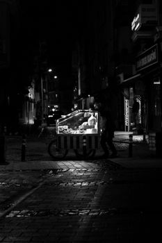 Black and white photo of a street vendor pushing a food cart in Çanakkale at night.