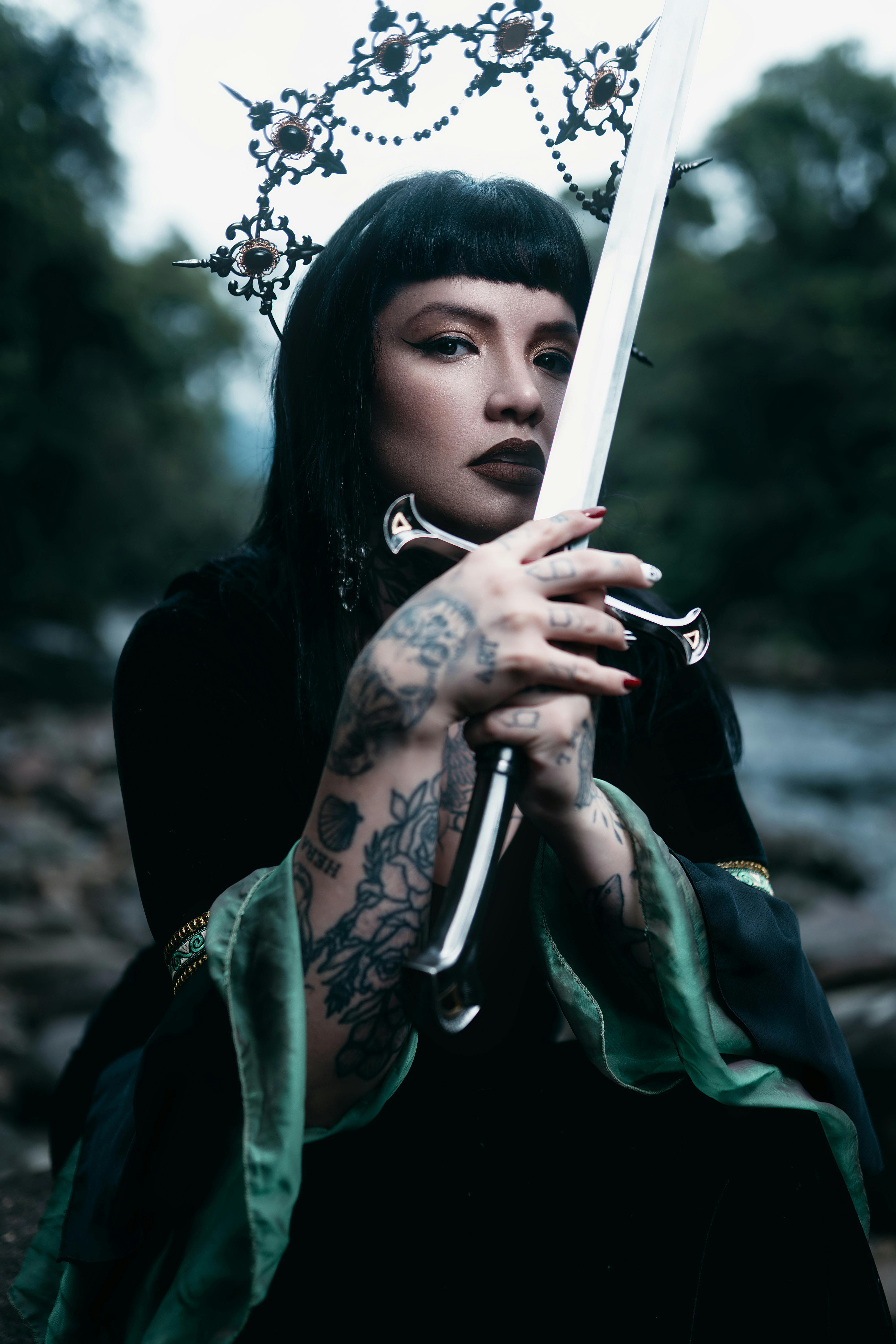Mystical portrait of tattooed woman holding ornate sword outdoors.