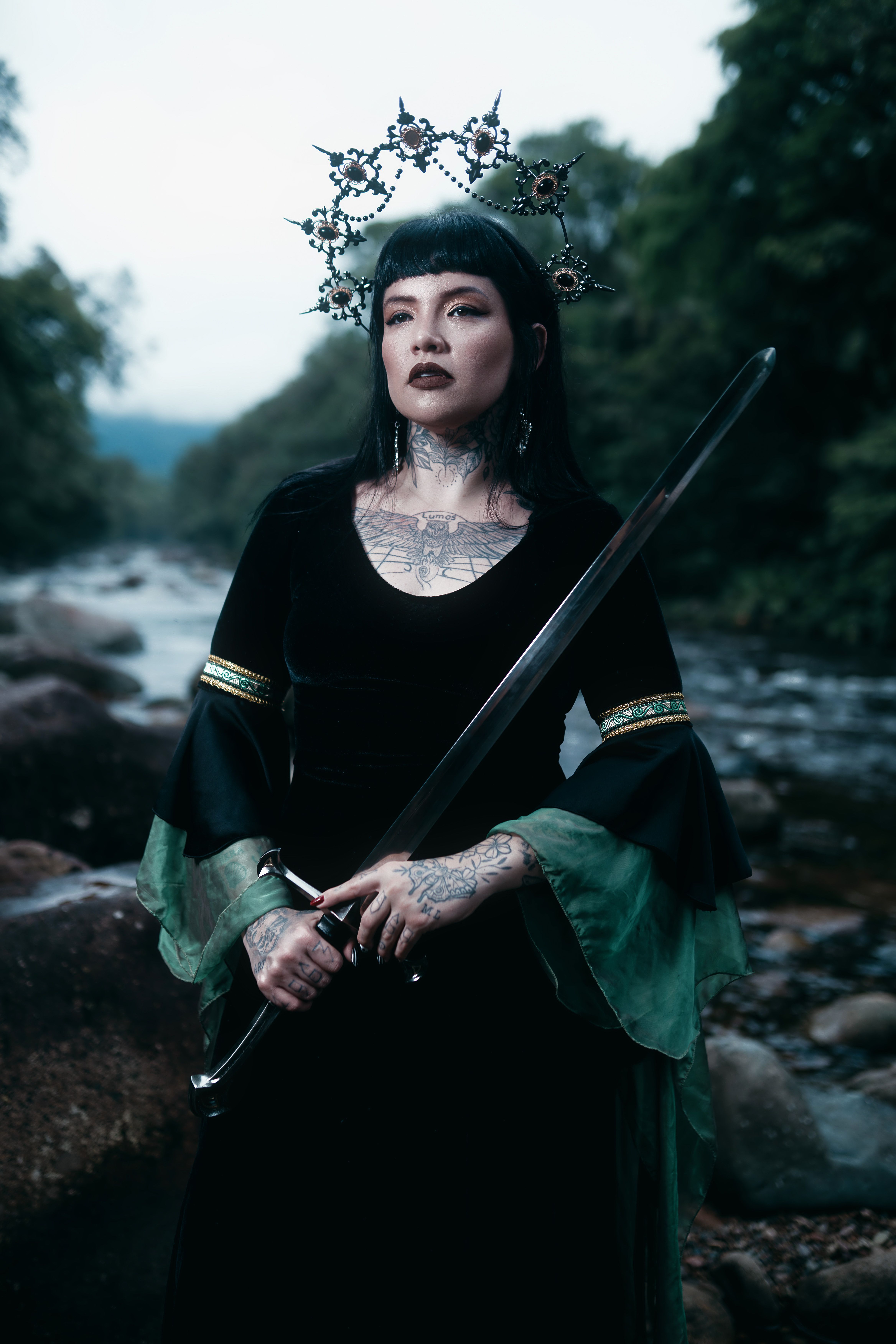 Tattooed woman in a medieval fantasy costume holds a sword by a scenic river.