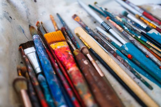 A variety of colorful, well-used artist paint brushes laid out on canvas cloth, showcasing creativity.