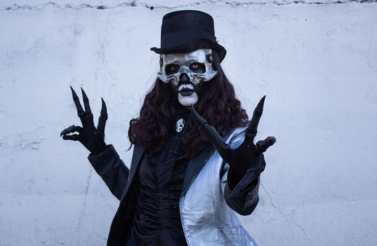 A person in a skeleton mask and top hat, dressed in dark formal attire with claw-like gloves, poses against a white wall.