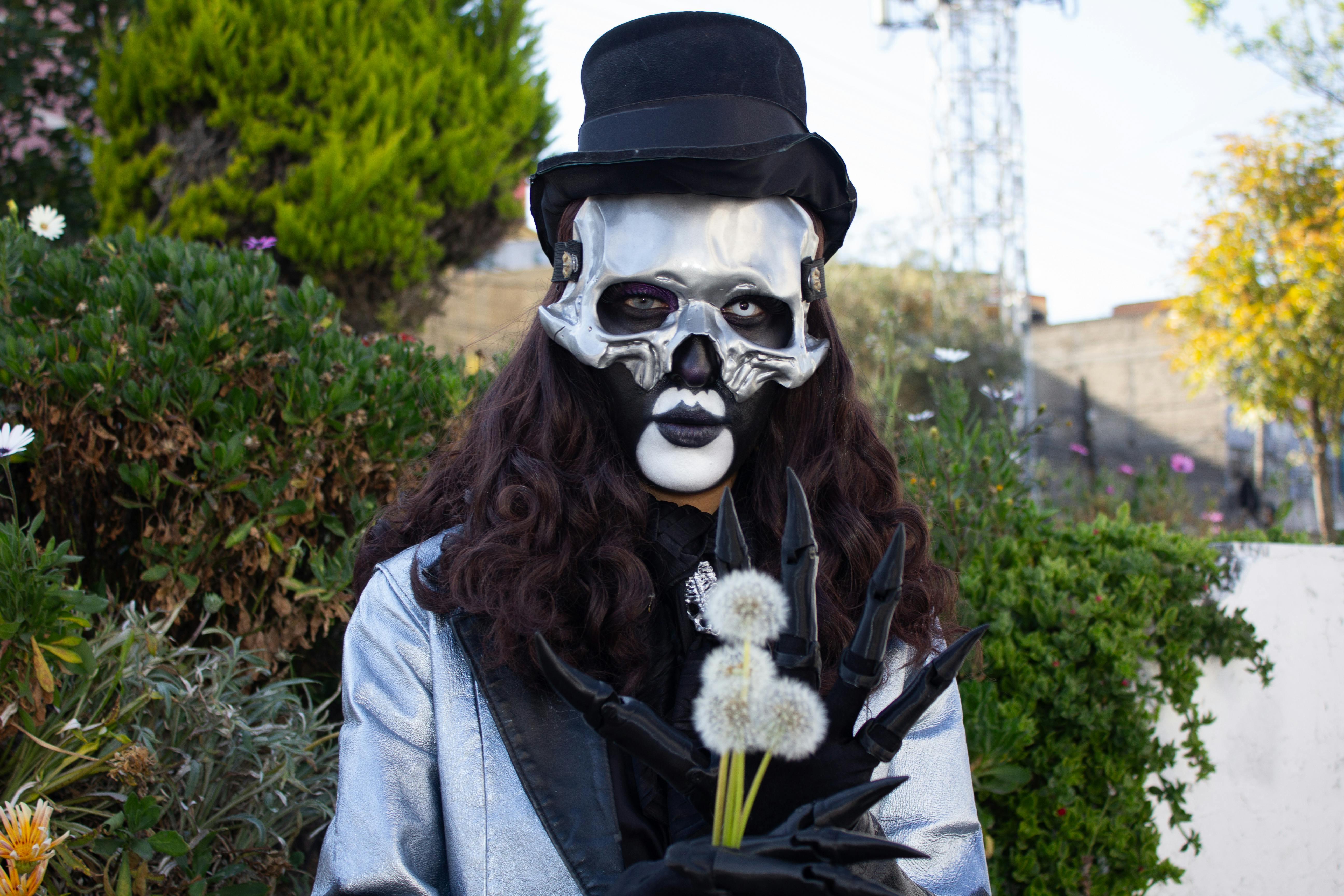 Gratis Una persona che indossa una maschera gotica e un cilindro tiene in mano dei denti di leone, creando una scena inquietante in un giardino all'aperto. Foto a disposizione
