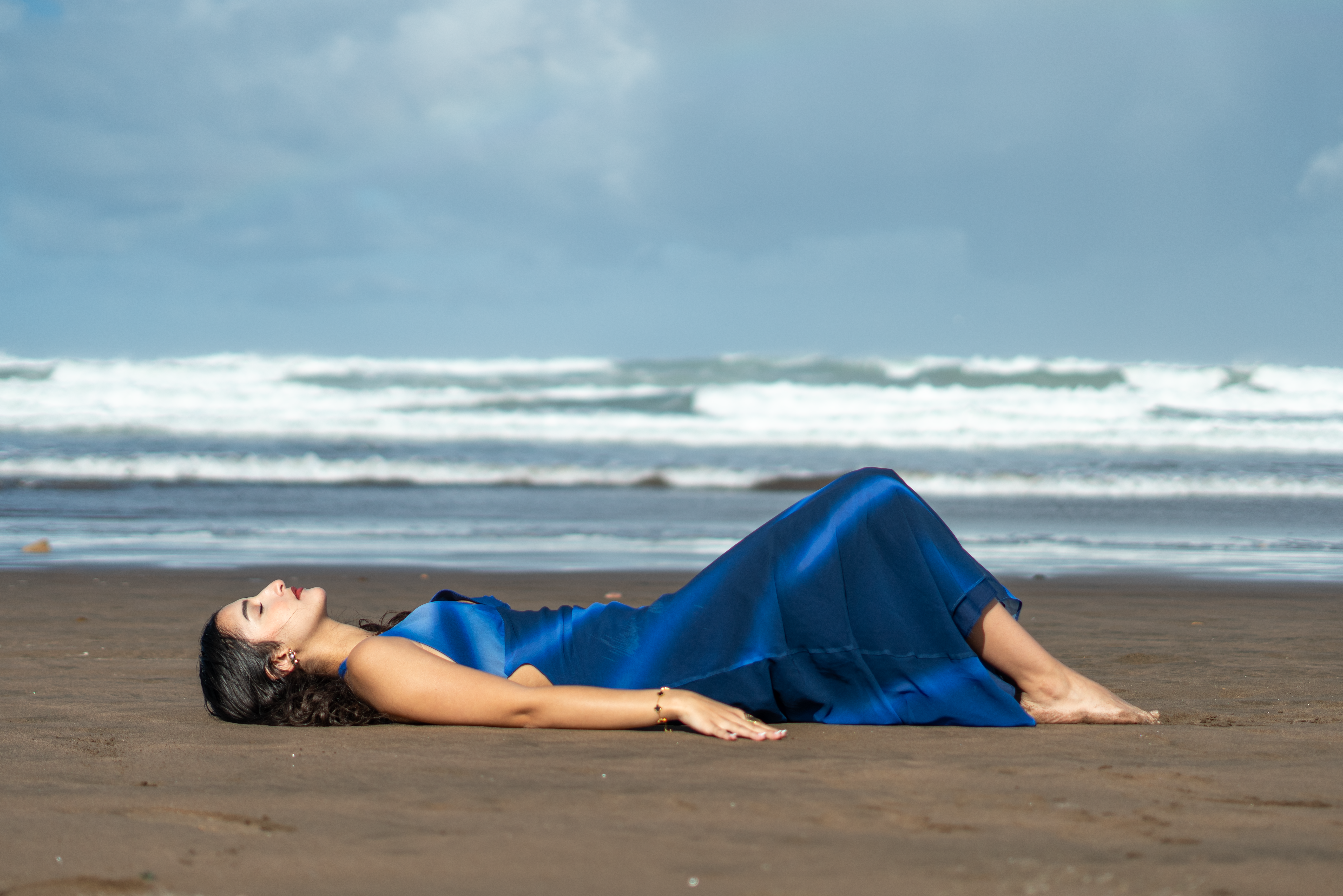 A woman in a blue dress lies relaxed on a sandy beach in Morocco with waves in the background.