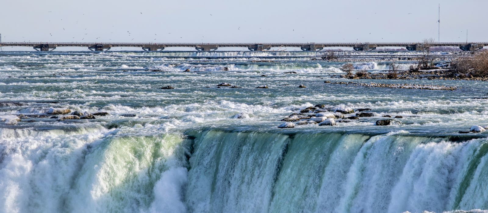 Scenic view of Niagara Falls with cascading water and a distant bridge under a clear sky.