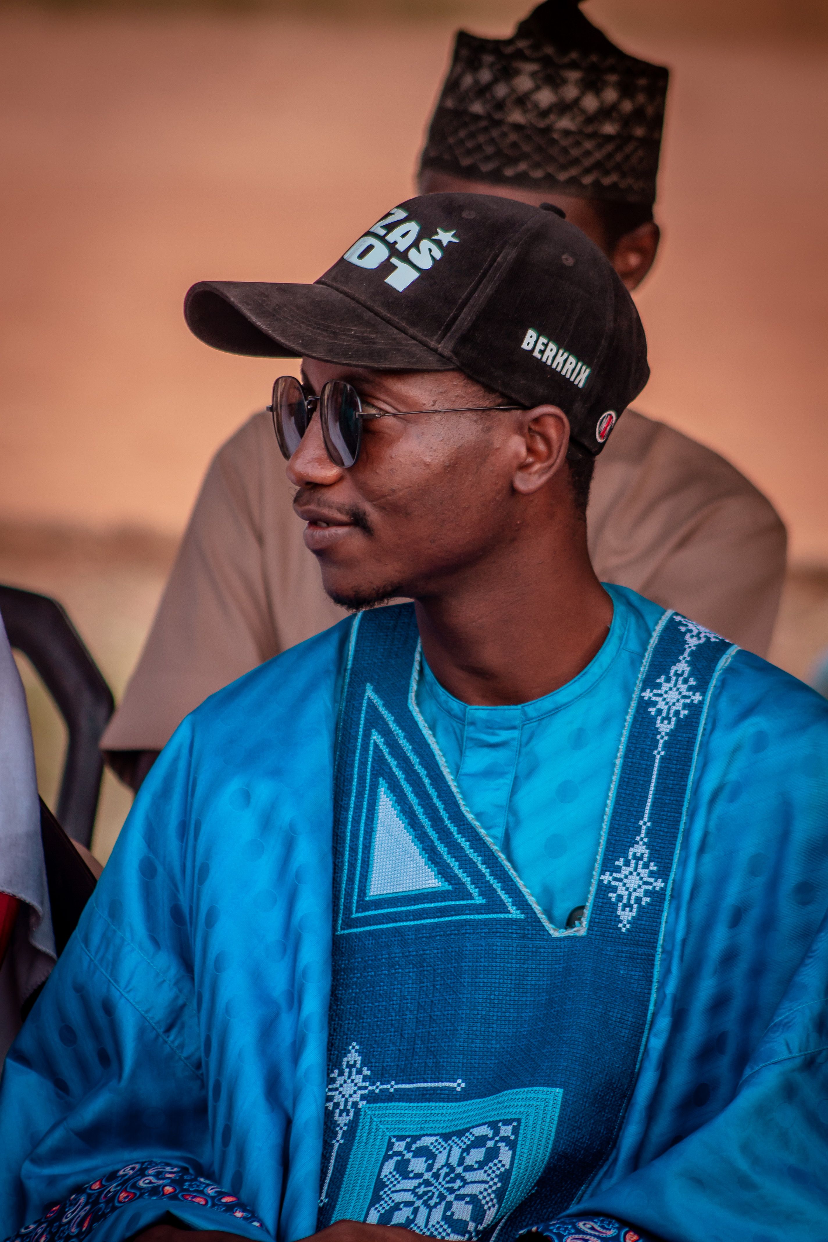 African man wearing traditional blue outfit and cap, seated outdoors.
