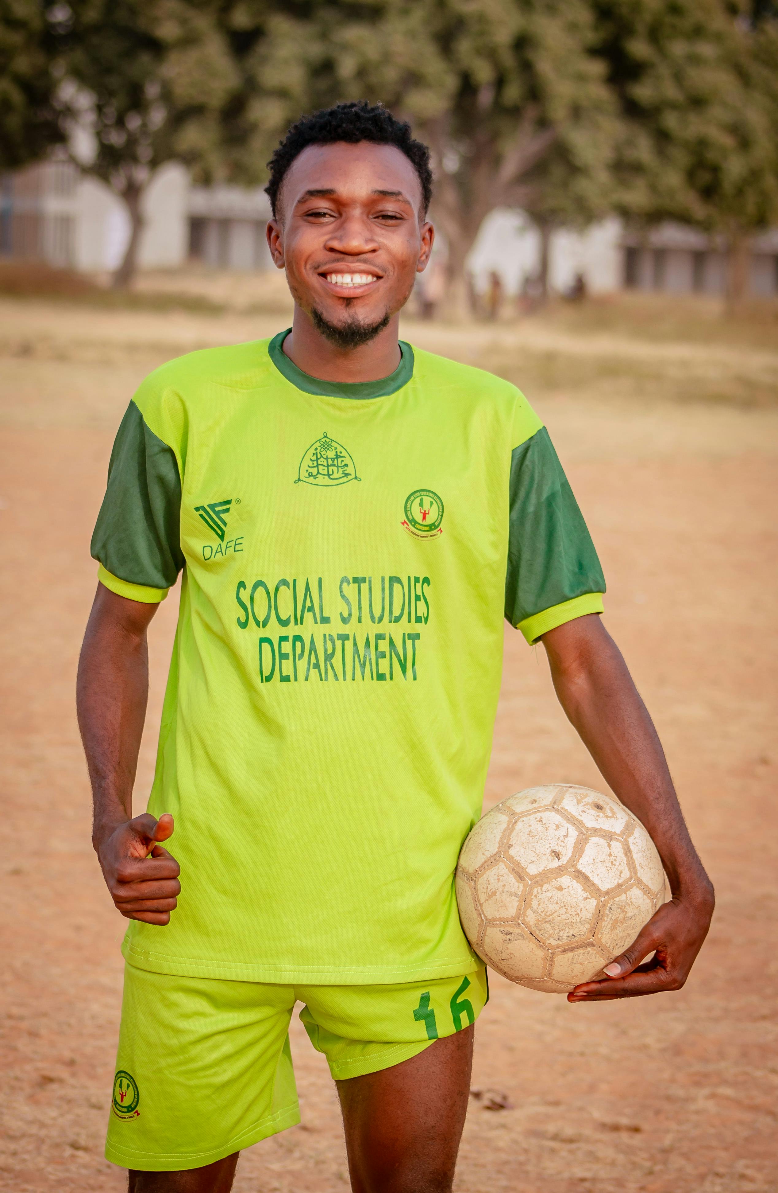Happy young man in green uniform holding a soccer ball, standing on an outdoor field.