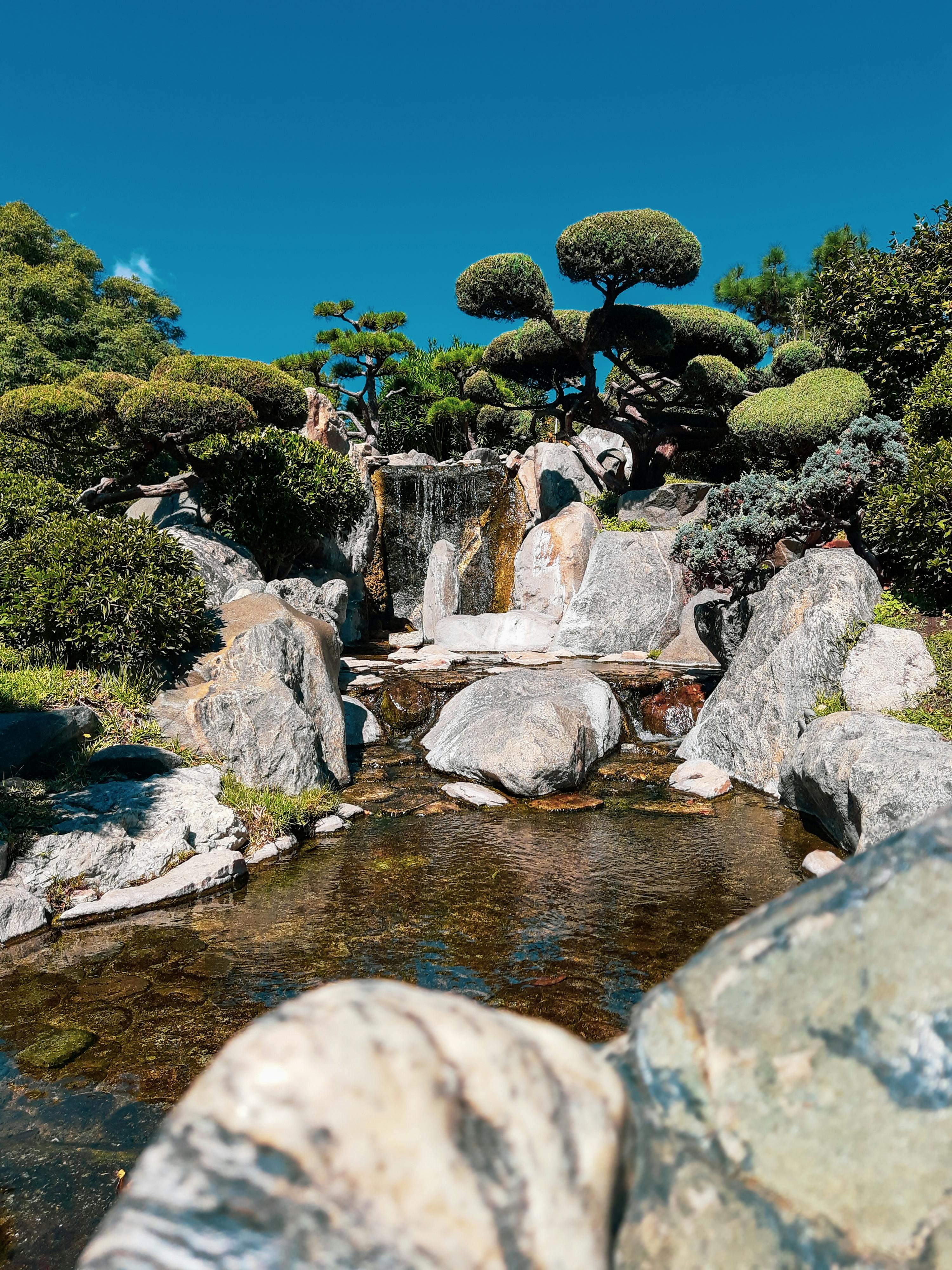 宁静的日式花园，有瀑布、岩石和修剪整齐的树木，头顶是湛蓝的天空。