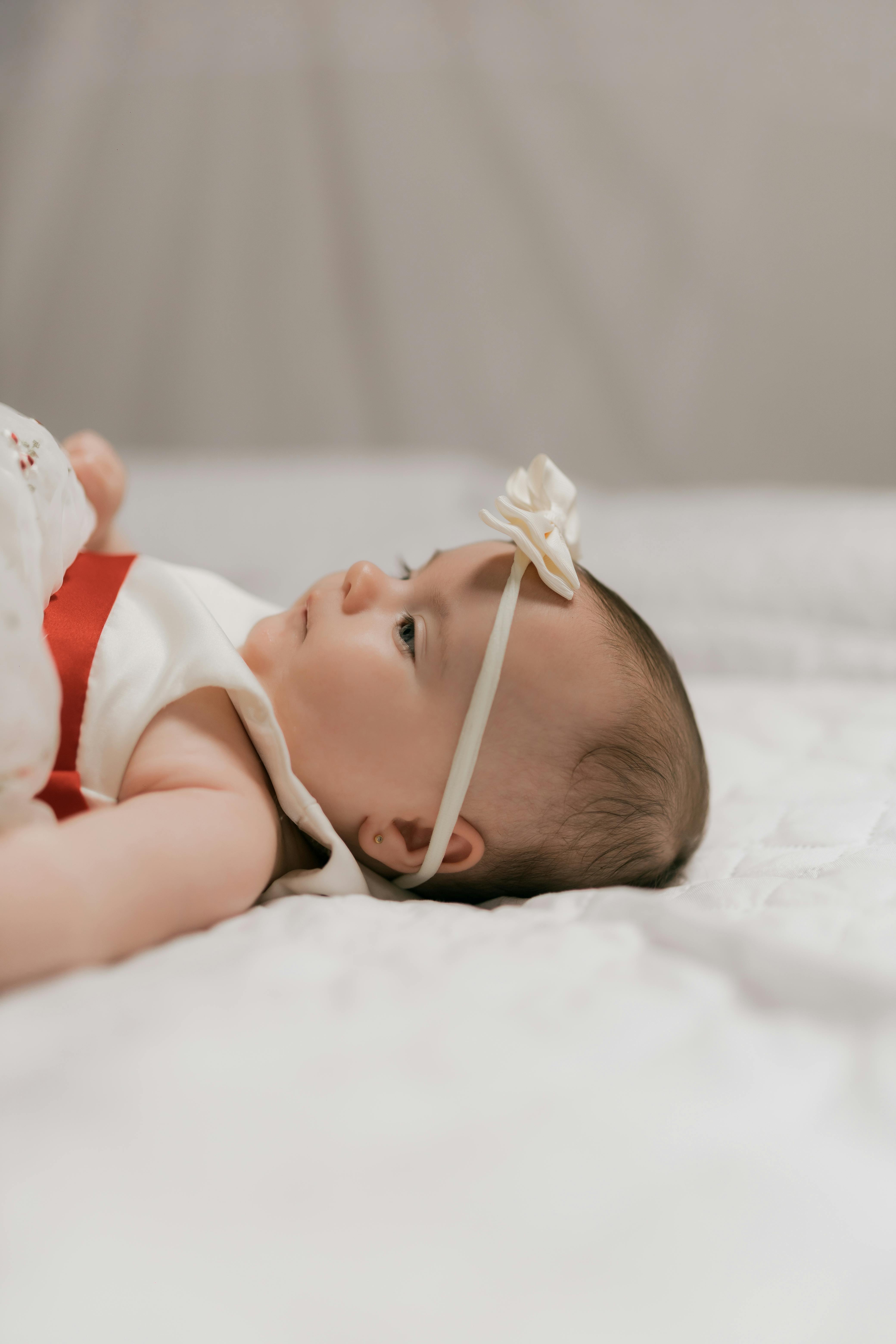 Cute baby wearing a white dress with a red accent and headband lying on a bed indoors.