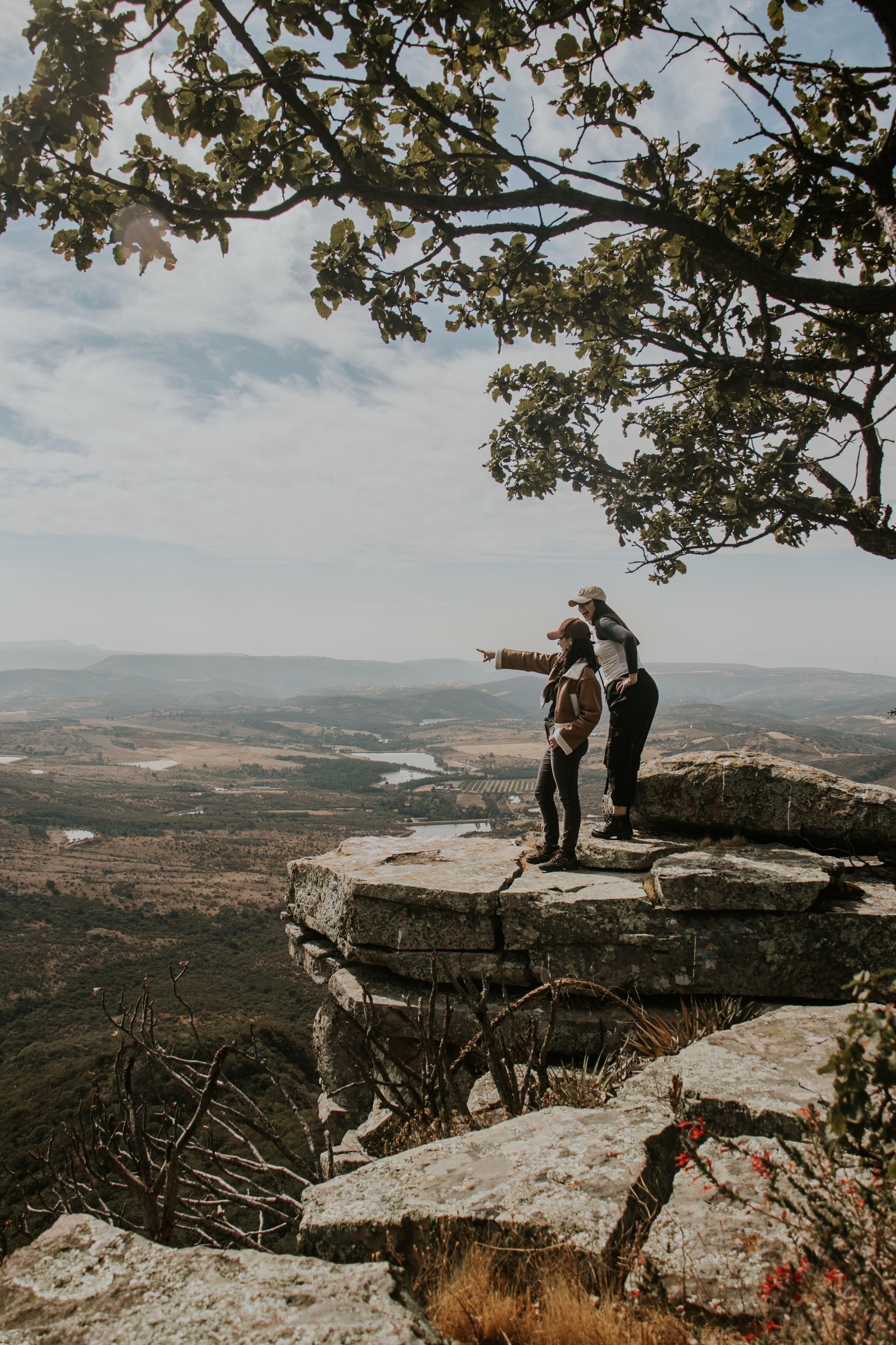 Two hikers admire an expansive valley view from a rocky cliff under a clear sky.