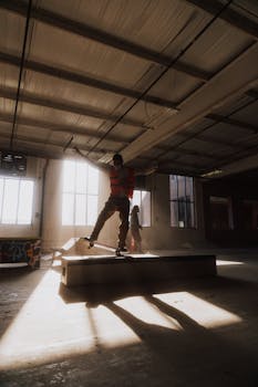 A skater performs a trick inside a sunlit, urban warehouse setting, casting long shadows.